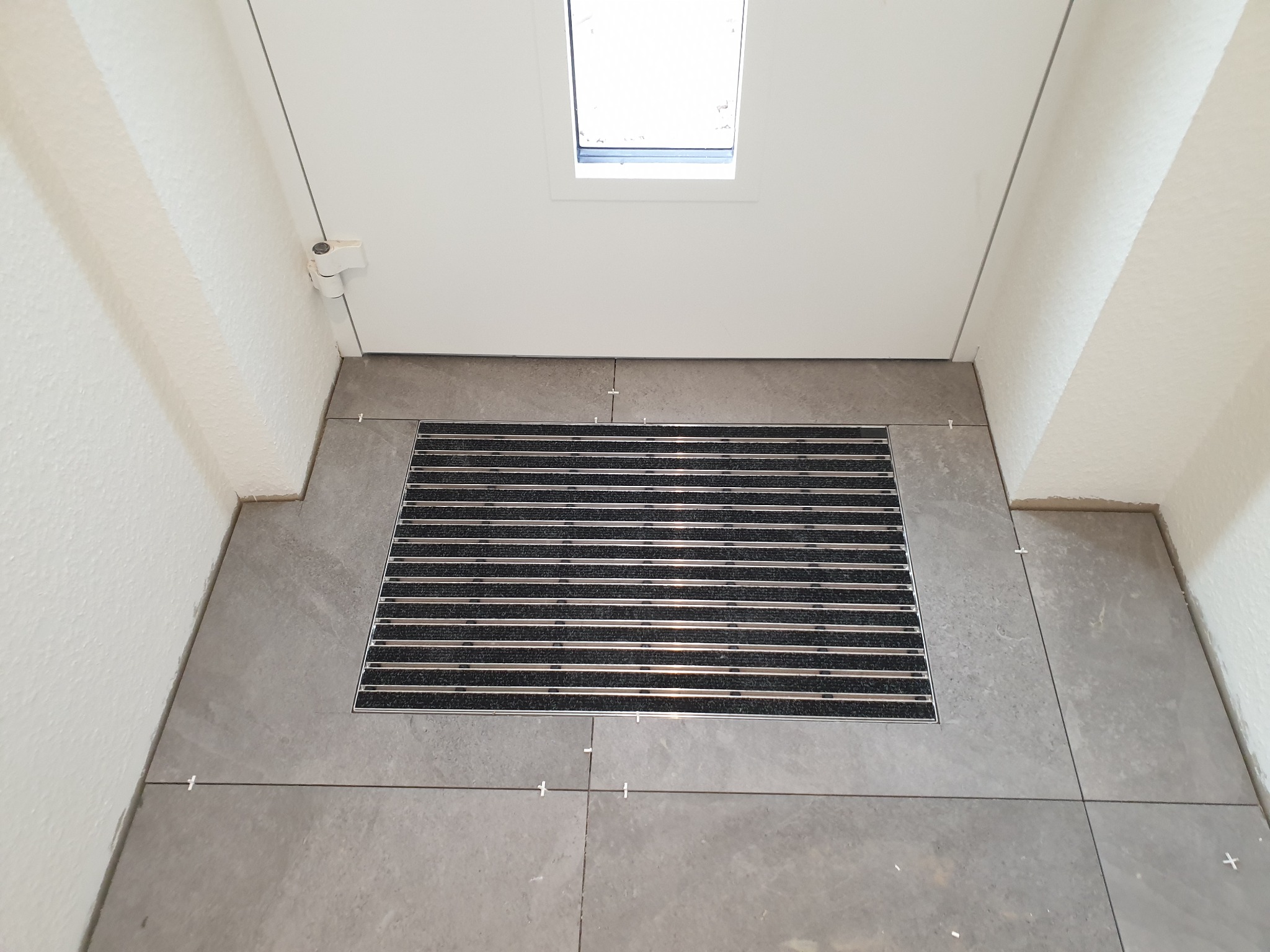 Montaż szarej, kwadratowej płytki podłogowej przy wejściu, z widoczną wycieraczką i krzyżykami dystansowymi.