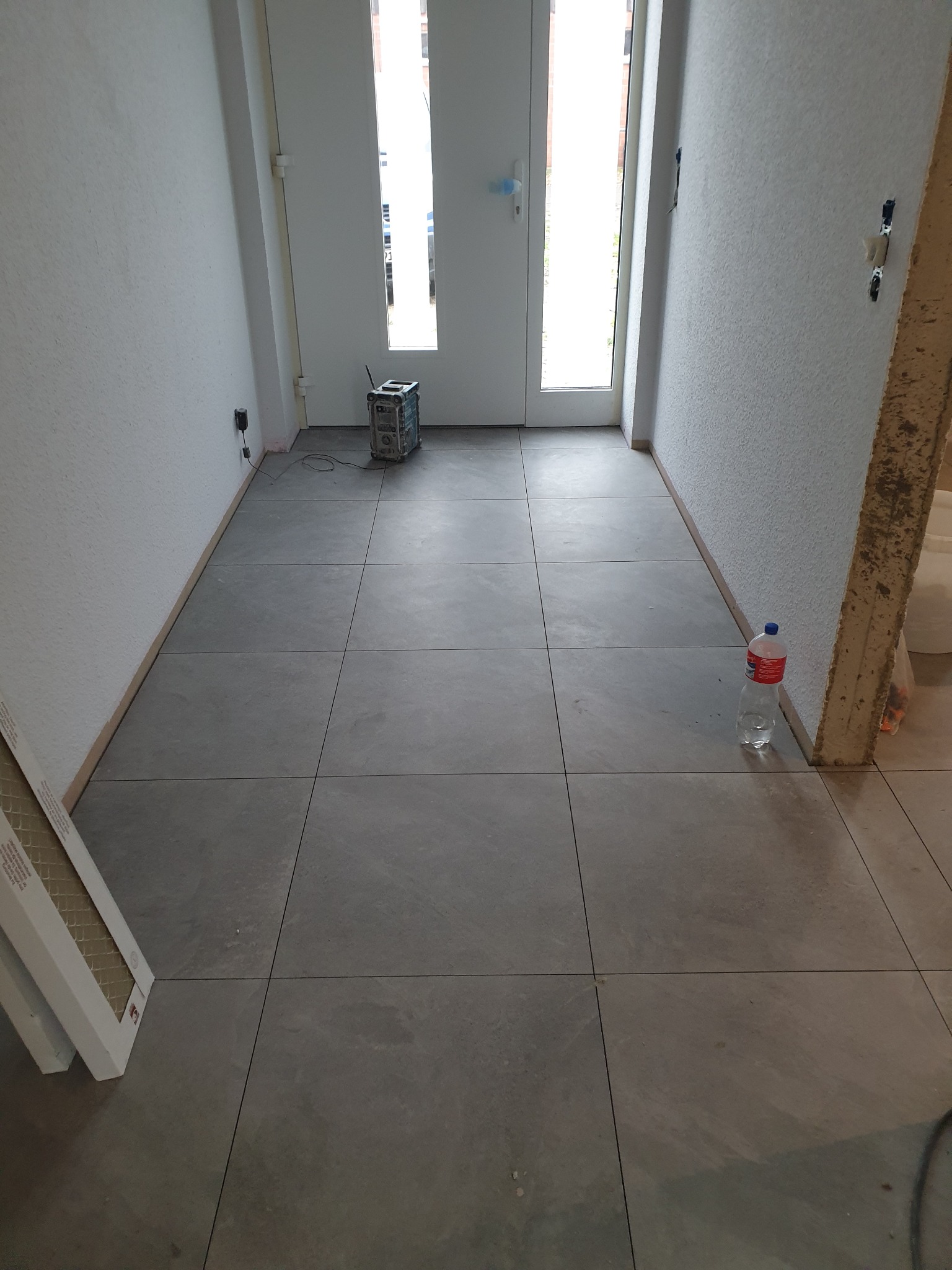 Ułożona szara, kwadratowa podłoga z płytek w korytarzu w Przytocznej, widoczna butelka wody i skrzynka narzędziowa. Częściowo wykończone ściany.