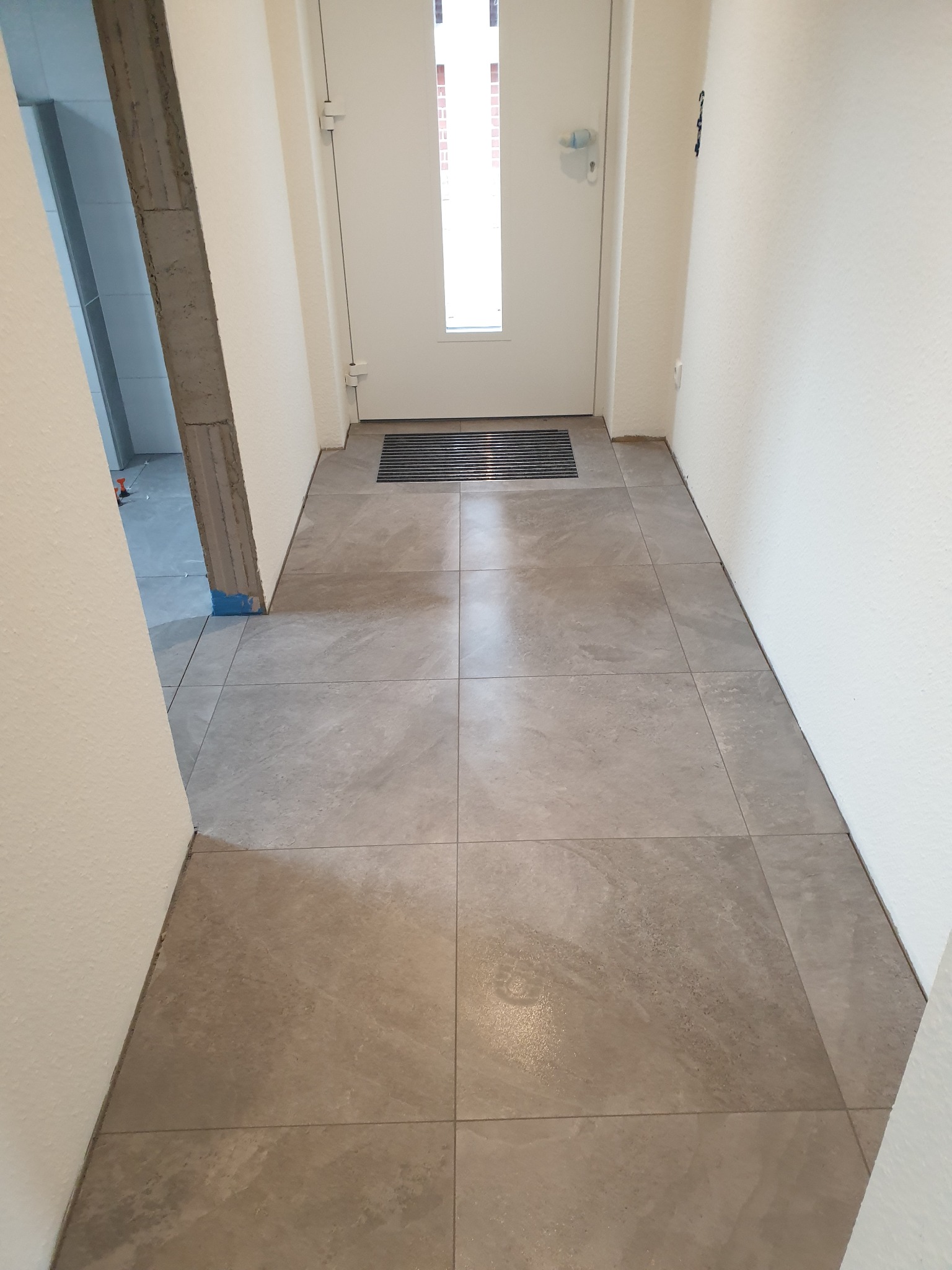 Korytarz wejściowy z nową, szarą podłogą z płytek gresowych imitujących kamień, widoczny fragment łazienki w trakcie prac wykończeniowych i białe ściany.