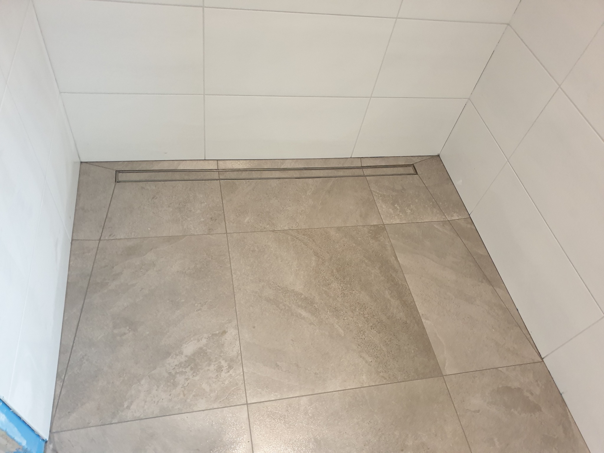 Ukończona kabina prysznicowa z odpływem liniowym, wyłożona beżowymi płytkami imitującymi kamień i białymi płytkami na ścianach.