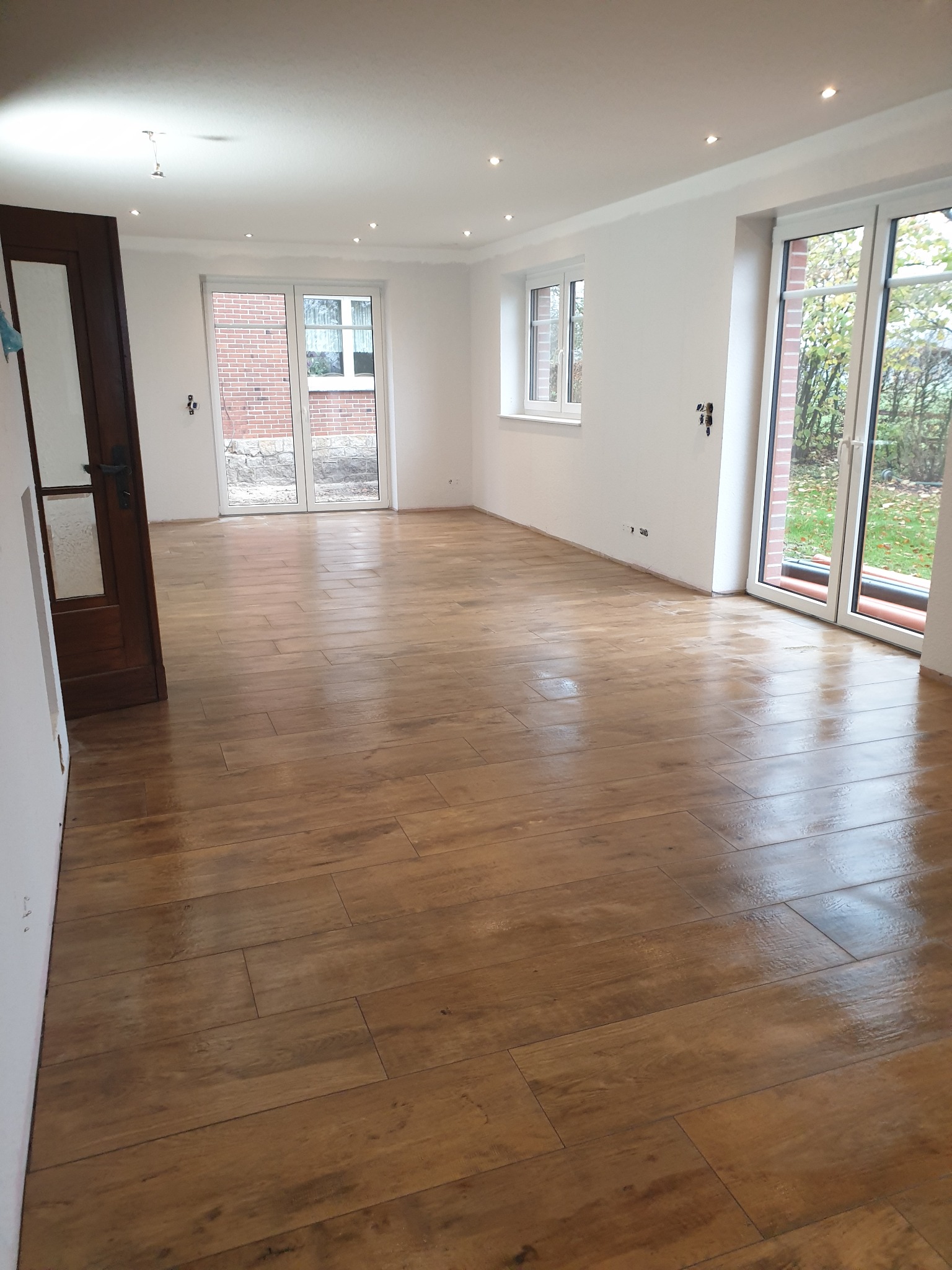 Pusta przestrzeń z nową, błyszczącą podłogą imitującą drewno, świeżo pomalowane białe ściany z widocznymi śladami po pracach wykończeniowych oraz otwarte drewniane drzwi i okna z widokiem na zewnątrz.
