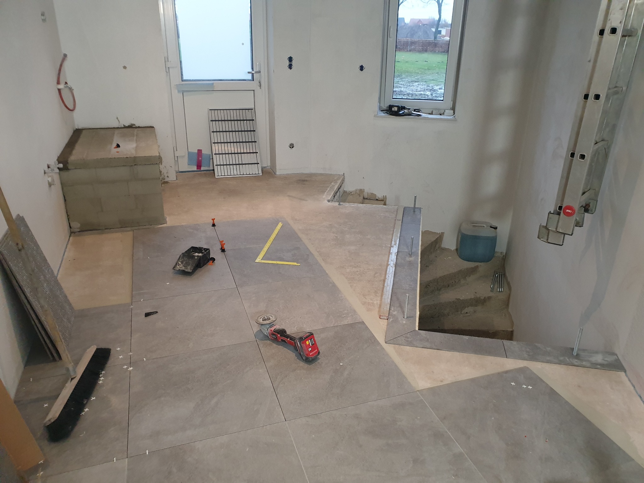 Układanie szarych płytek podłogowych w pomieszczeniu w trakcie remontu, widoczna szlifierka kątowa, poziomica, krzyżyki dystansowe i szczotka do fugowania.