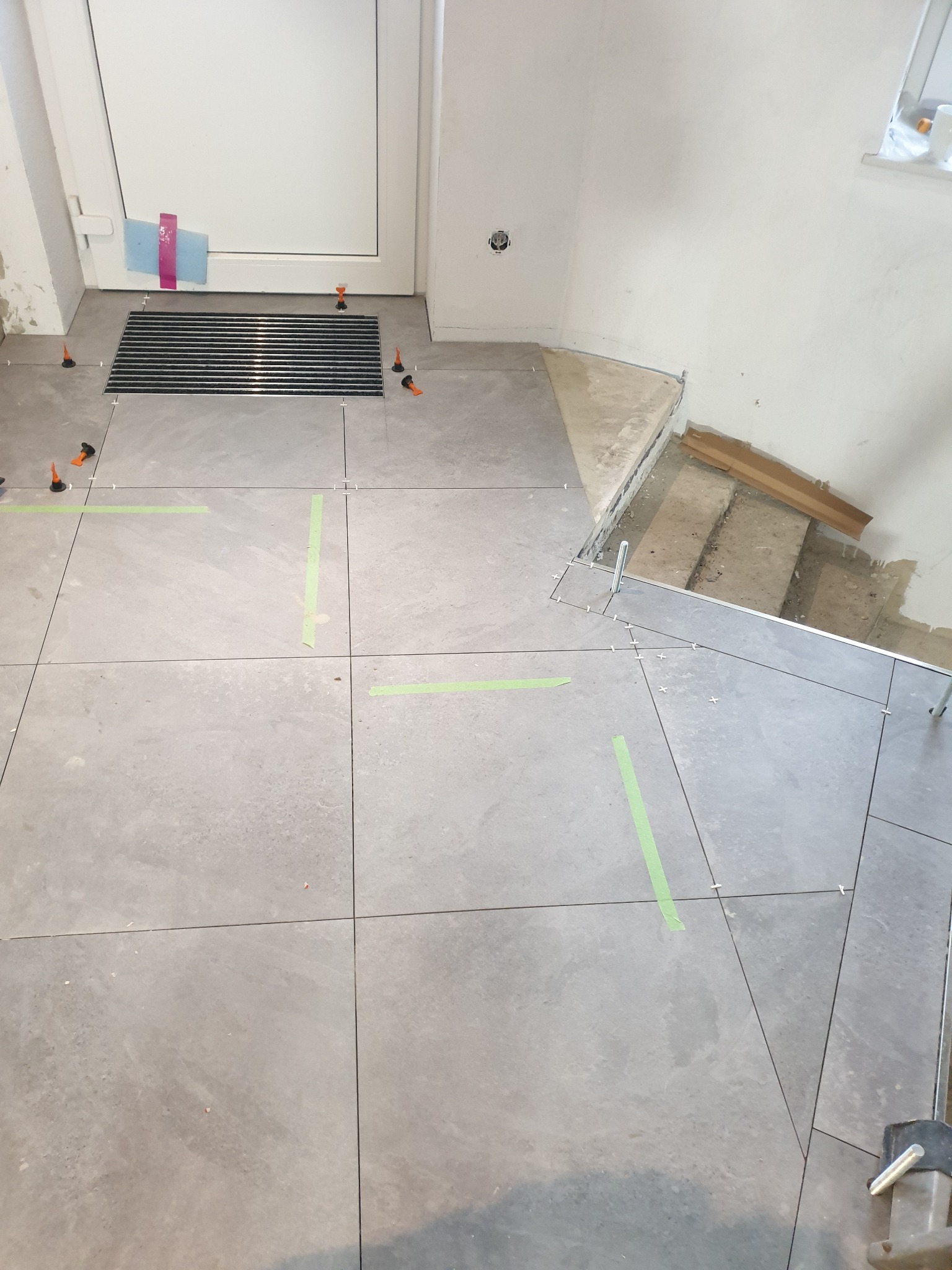 Układanie szarych płytek podłogowych w przedpokoju, widoczne poziomice, krzyżyki dystansowe i taśma malarska, fragment schodów w tle.
