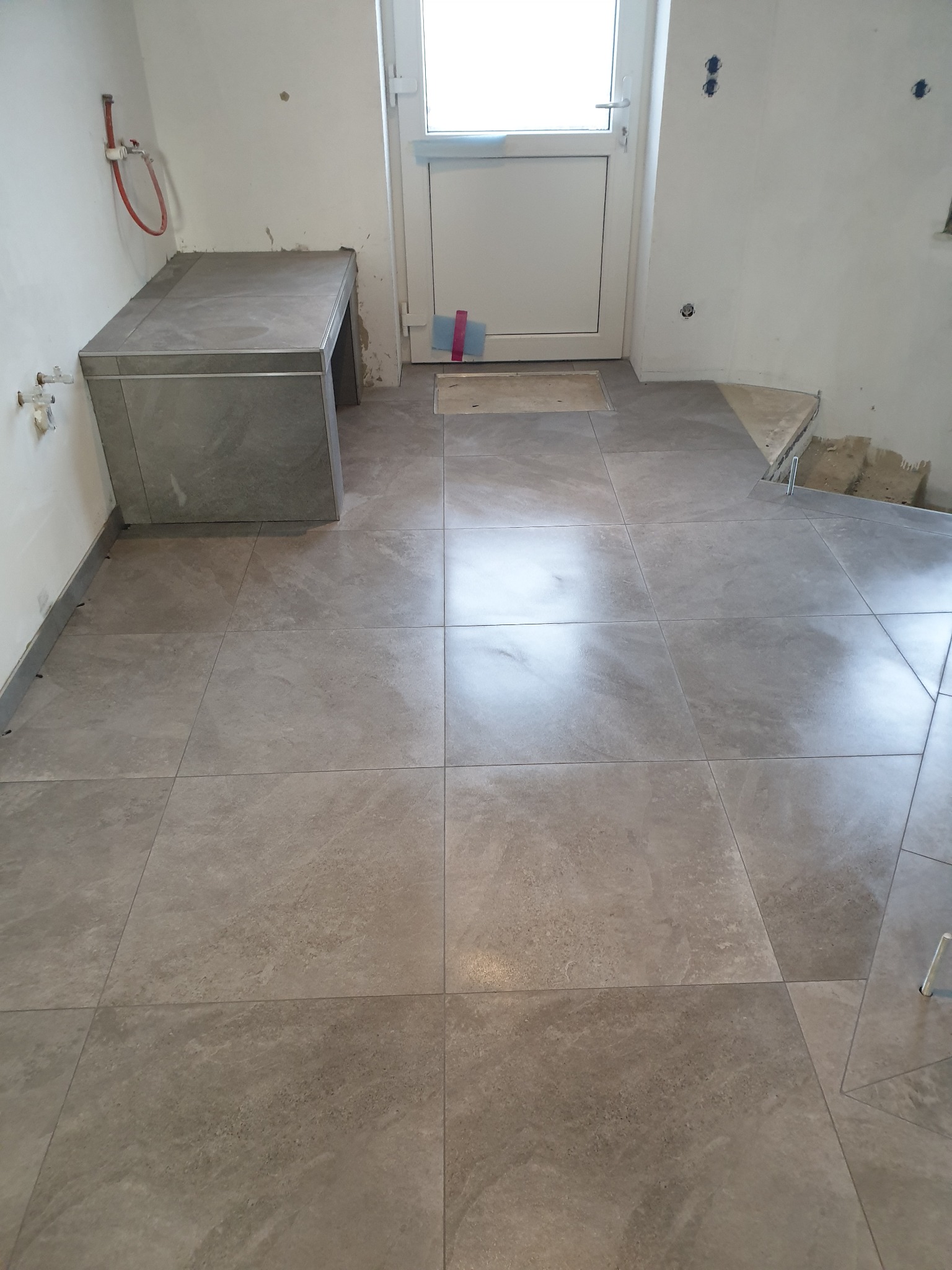 Ułożona szara terakota w łazience w trakcie remontu, widoczne niedokończone ściany i instalacje hydrauliczne, z fragmentem zabudowy pod umywalkę wykończonej tymi samymi płytkami.