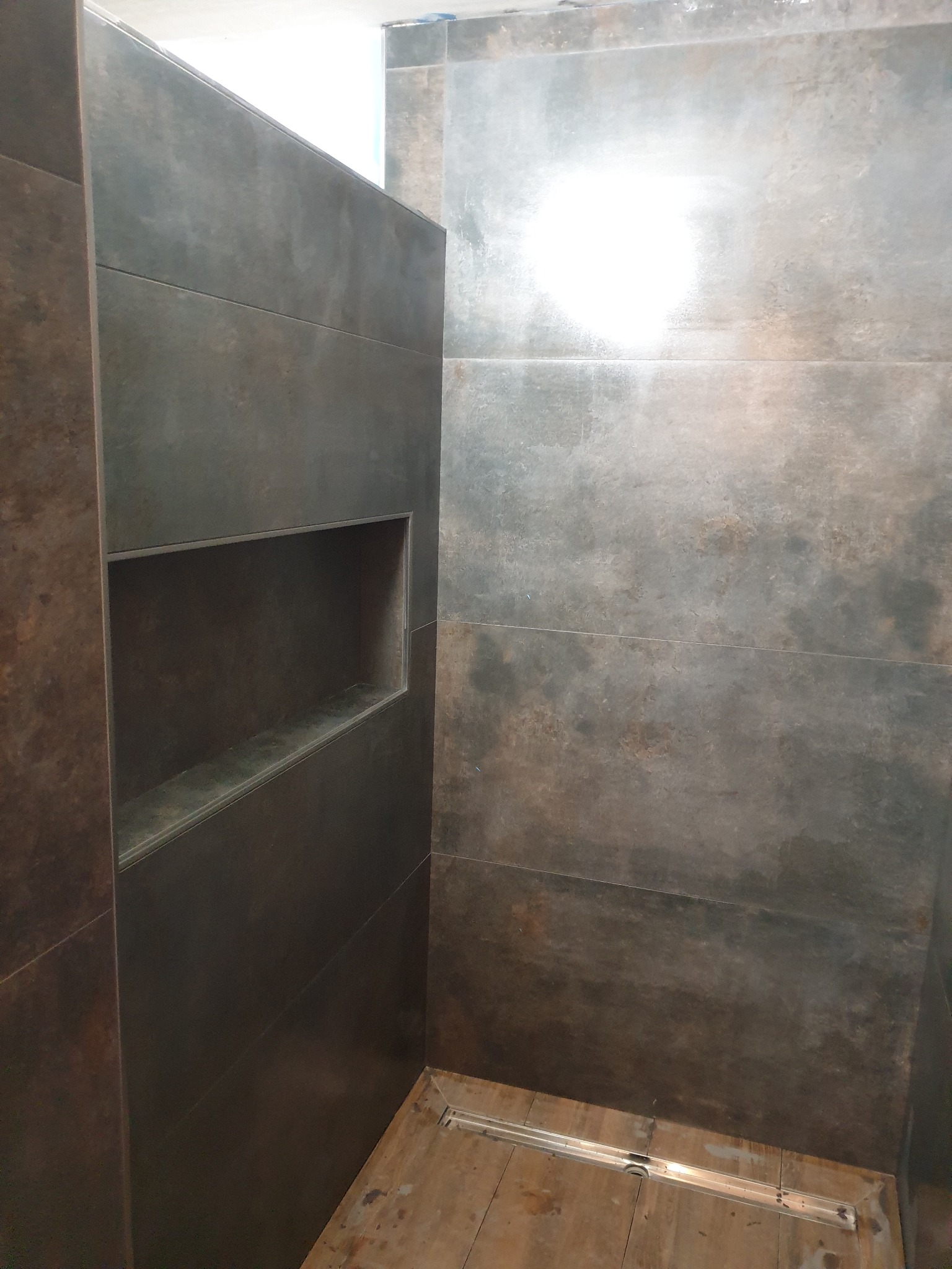 Wykończona kabina prysznicowa z ciemnymi, płytkami imitującymi metal, wbudowaną półką i odpływem liniowym w podłodze.