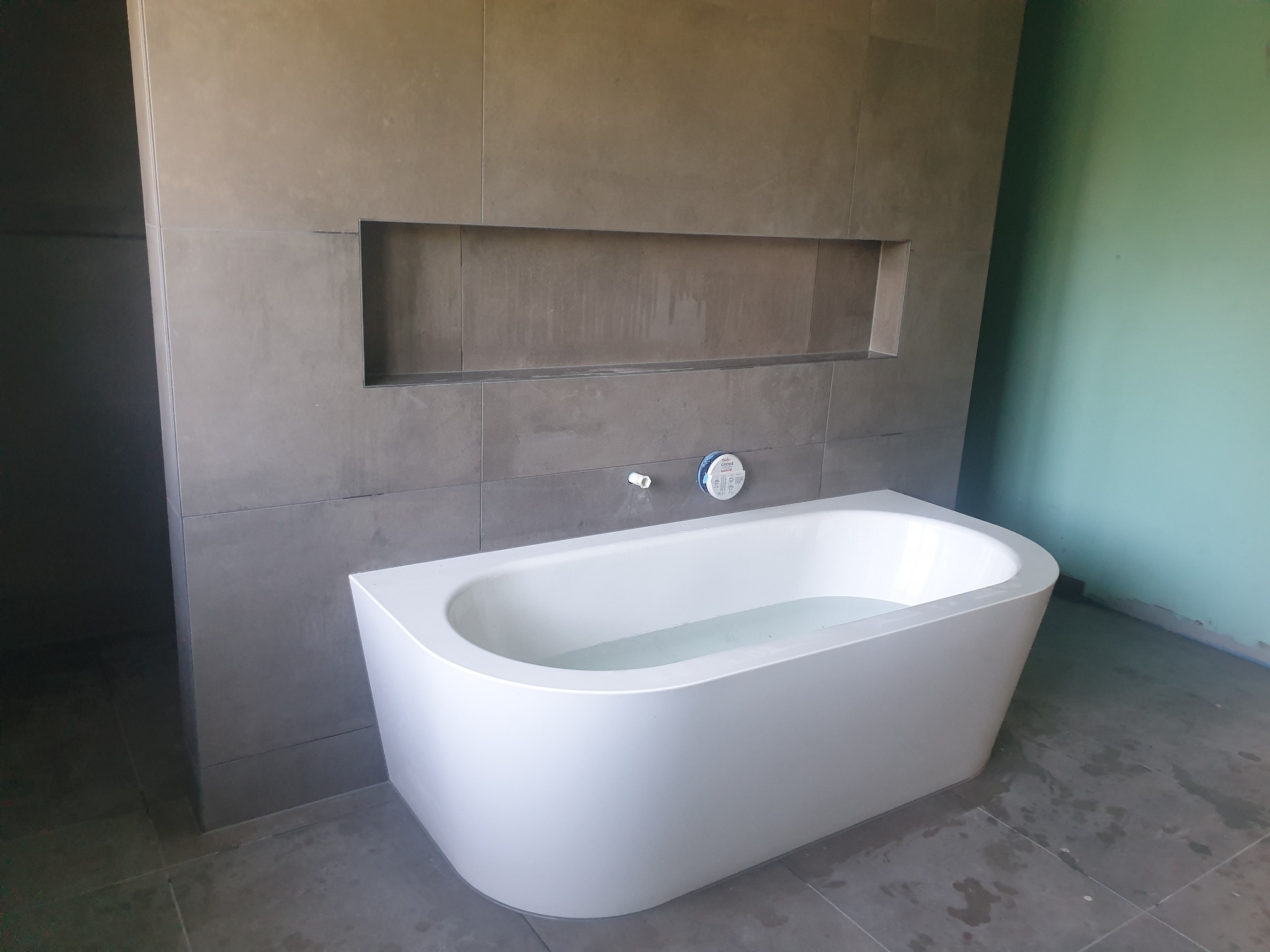 Nowoczesna, wolnostojąca wanna wypełniona wodą, na tle szarych płytek z wnęką w ścianie i fragmentem zielonej ściany, widoczne ślady po remoncie.