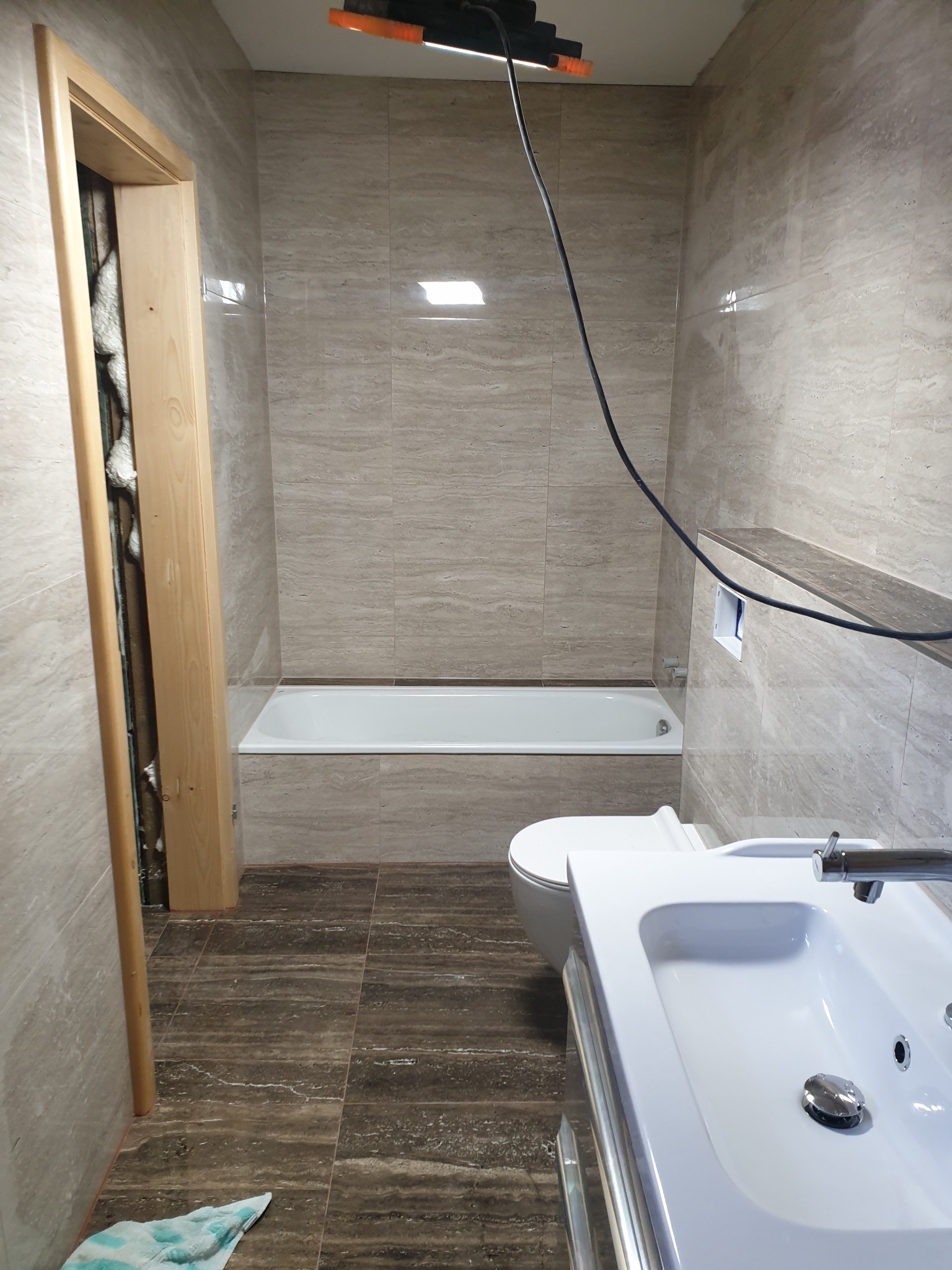 Wnętrze łazienki w trakcie prac wykończeniowych, widoczna nowa armatura, płytki imitujące trawertyn na ścianach, ciemne płytki na podłodze, ościeżnica drzwiowa z pianką montażową, lampa robocza...