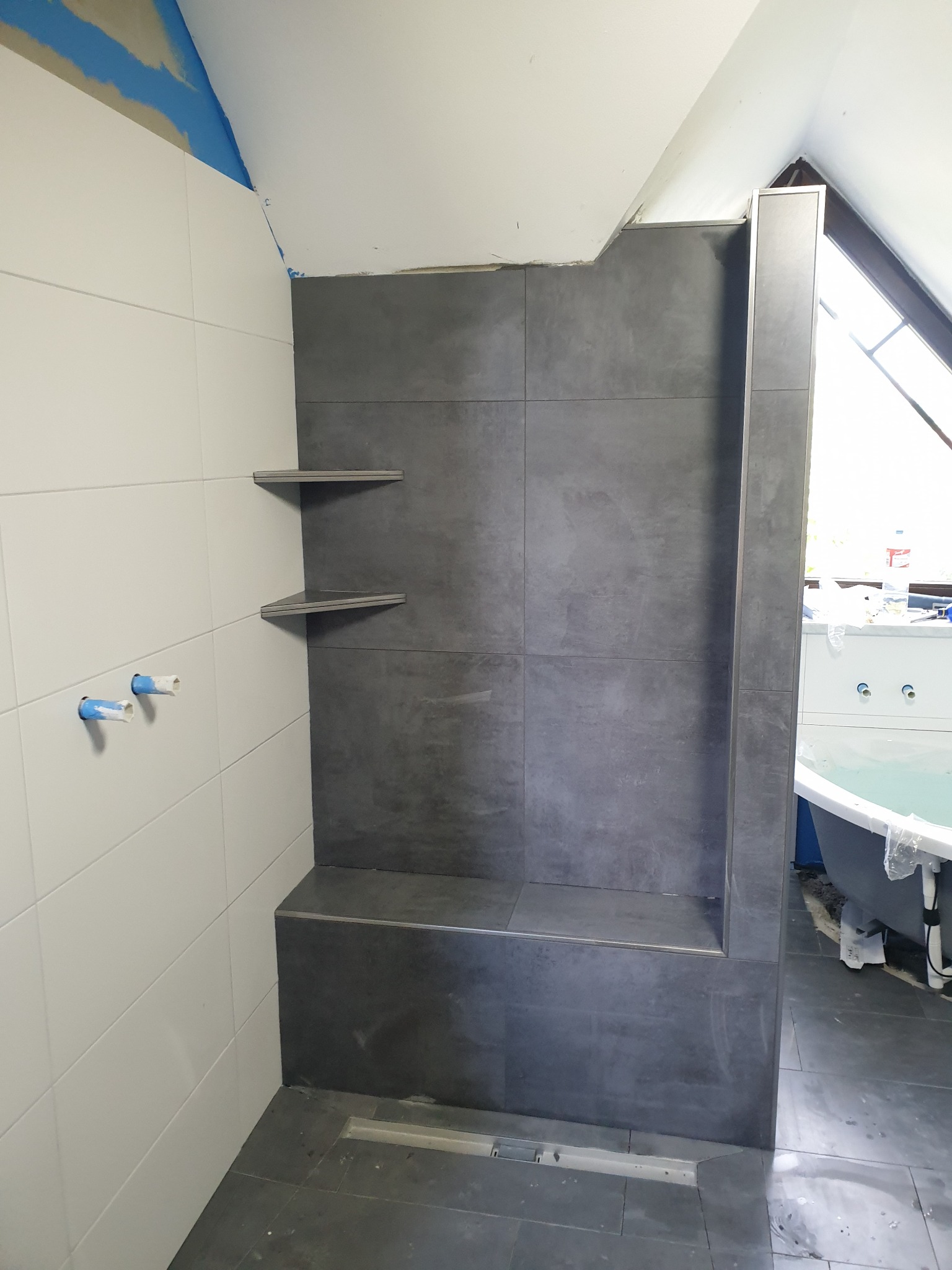 Częściowo wykończona łazienka na poddaszu z szarymi płytkami, wnęką prysznicową z siedziskiem i półkami narożnymi, białe płytki na ścianie z widocznymi rurami oraz odpływ liniowy w podłodze.