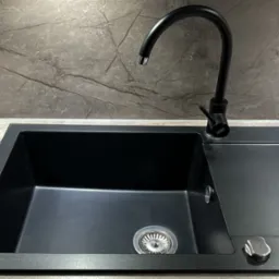 Czarny, prostokątny zlewozmywak granitowy z ociekaczem i czarną baterią na tle płytek imitujących marmur.