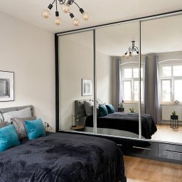 Studio Kuchni&Salon Meblowy Design Plus - Sypialnia z łóżkiem z szarym zagłówkiem i turkusowymi poduszkami, duża szafa przesuwna z lustrami odbija wnętrze pokoju, na suficie lampa z żarówkami Edisona.