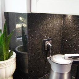 Studio Kuchni&Salon Meblowy Design Plus - Fragment blatu kuchennego z czarnego kamienia z połyskującymi drobinkami, gniazdko elektryczne z wtyczką, czajnik elektryczny oraz doniczka z zieloną rośliną przy oknie.