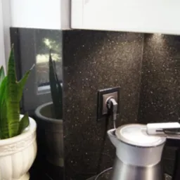 Fragment blatu kuchennego z czarnego kamienia z połyskującymi drobinkami, gniazdko elektryczne z wtyczką, czajnik elektryczny oraz doniczka z zieloną rośliną przy oknie.