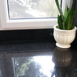 Studio Kuchni&Salon Meblowy Design Plus - Błyszczący, czarny blat z drobnymi, białymi plamkami, ustawiony przy oknie z widokiem na zieleń, ozdobiony doniczką z sansewierią i fragmentem marmurowego moździerza.