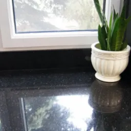 Błyszczący, czarny blat z drobnymi, białymi plamkami, ustawiony przy oknie z widokiem na zieleń, ozdobiony doniczką z sansewierią i fragmentem marmurowego moździerza.