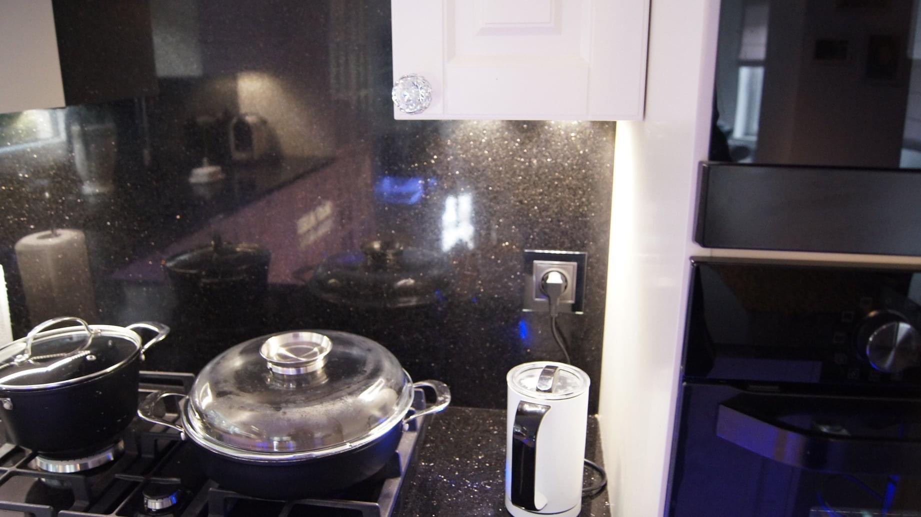 Fragment kuchni z czarnym blatem o fakturze galaktyki, na którym stoją dwa garnki na płycie gazowej i biały czajnik elektryczny, obok widać białą szafkę z ozdobnym, kryształowym uchwytem...