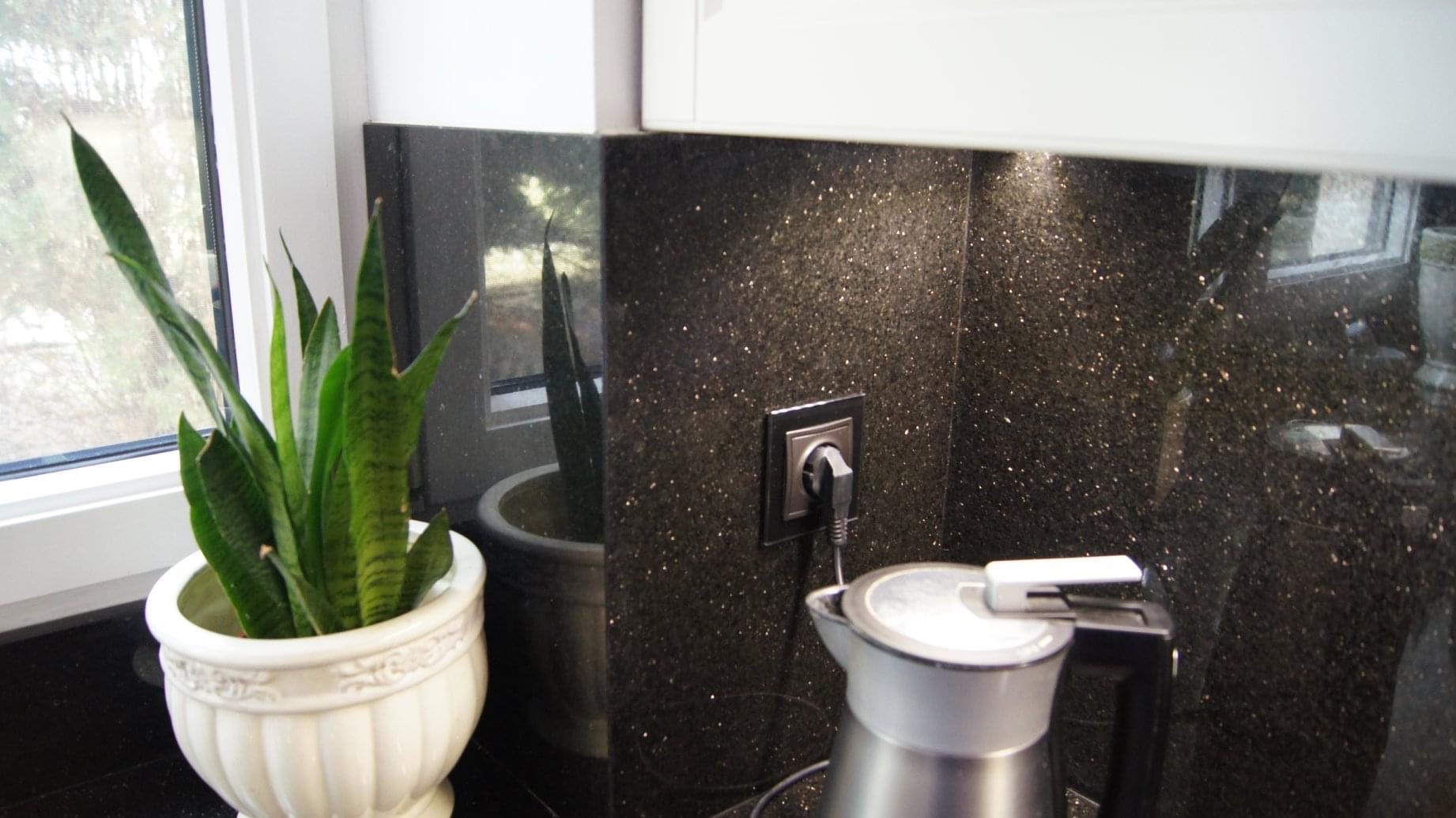 Fragment blatu kuchennego z czarnego kamienia z połyskującymi drobinkami, gniazdko elektryczne z wtyczką, czajnik elektryczny oraz doniczka z zieloną rośliną przy oknie.