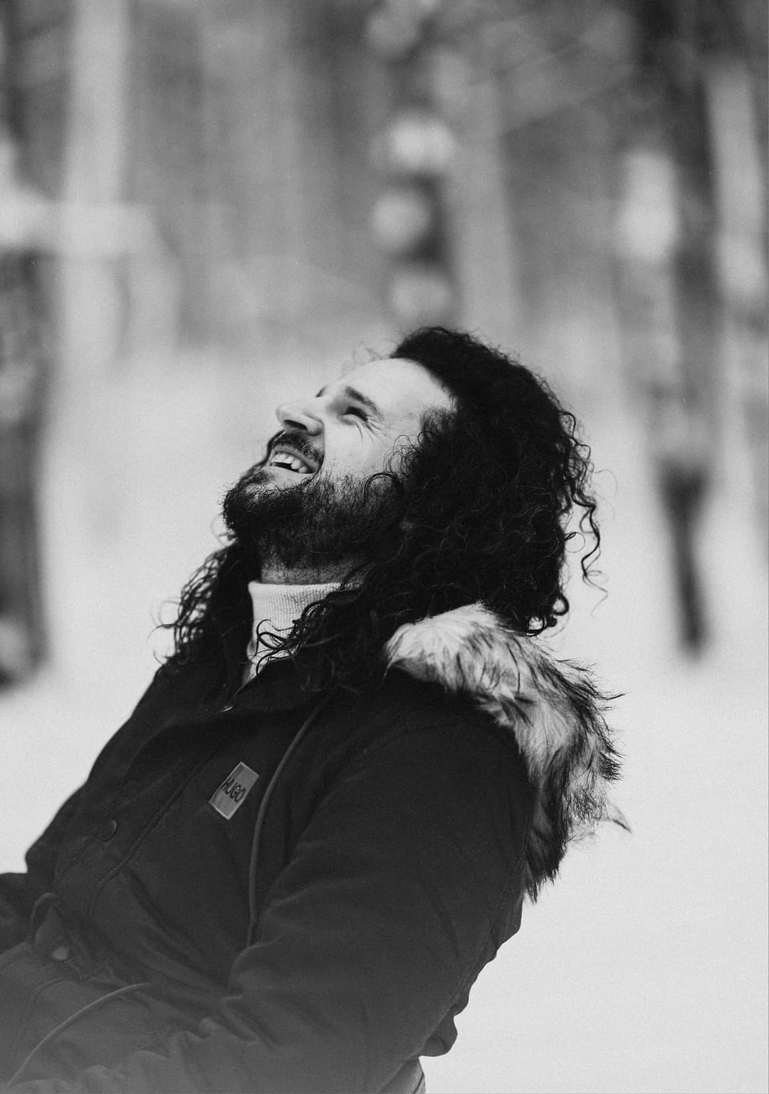 Czarno-białe zdjęcie uśmiechniętego mężczyzny z brodą, spoglądającego w górę na tle zimowego krajobrazu, ubrany w kurtkę z futrzanym kapturem.