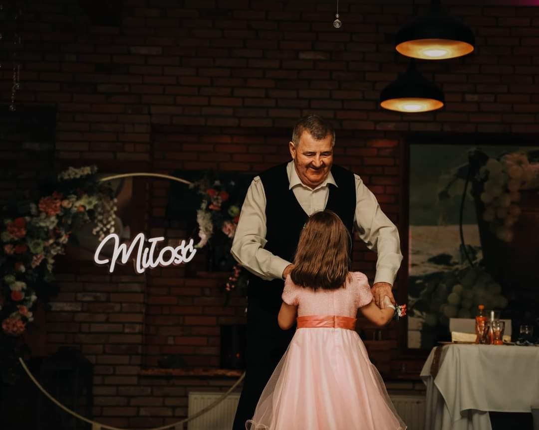 Dziadek w czarnej kamizelce tańczy z wnuczką w różowej sukience na tle ceglanej ściany z neonowym napisem 'Miłość' i kwiatową dekoracją podczas uroczystości.
