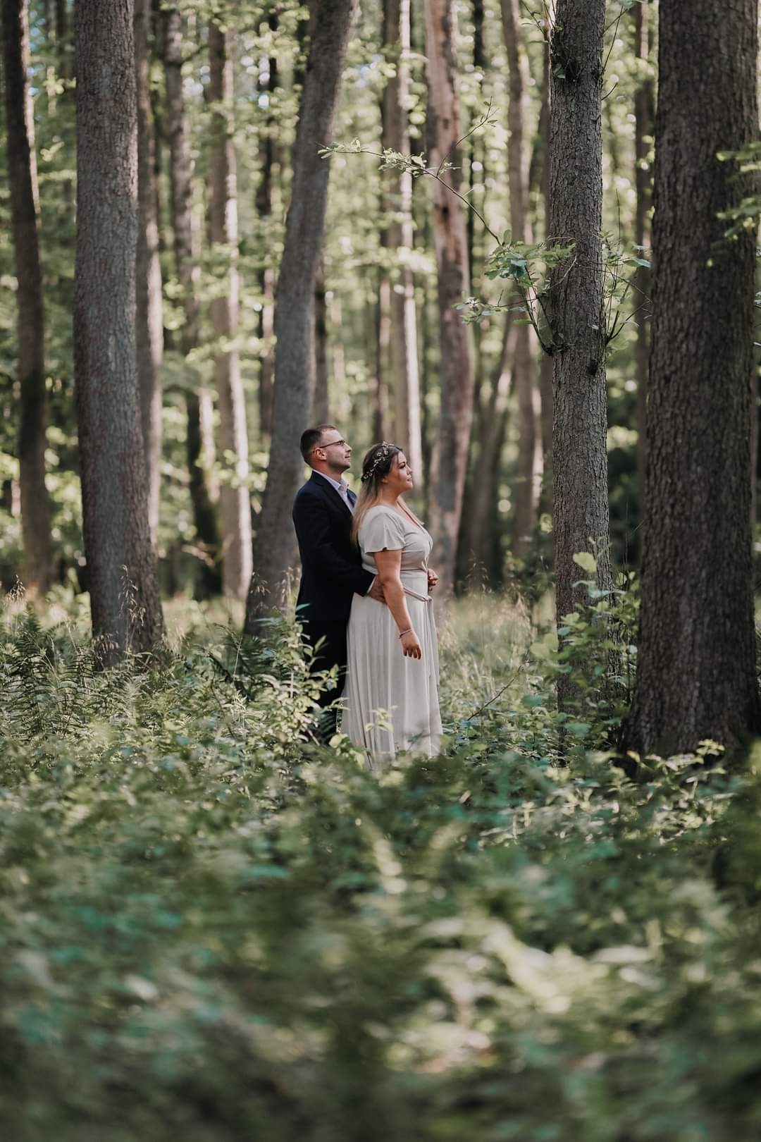 Para w strojach ślubnych stoi w lesie, otoczona zieloną roślinnością, oboje patrzą w górę, mężczyzna w ciemnym garniturze obejmuje kobietę w długiej, jasnej sukni.