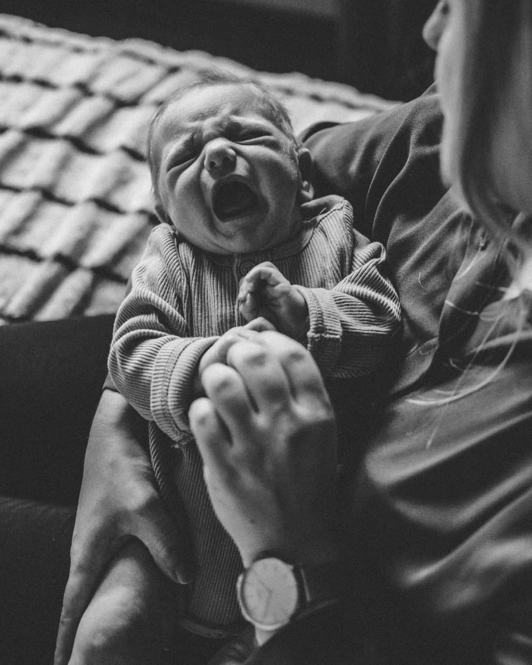 Czarne i białe zdjęcie płaczącego noworodka w prążkowanym ubranku trzymanego przez osobę z zegarkiem na ręku, z widocznym fragmentem wzorzystej pościeli w tle.