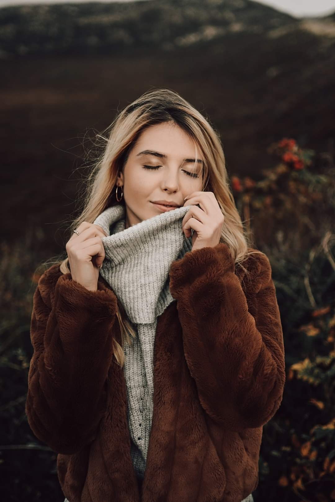 Portret kobiety z zamkniętymi oczami, w brązowym futrze i szarym swetrze, delikatnie dotykającej kołnierza, z rozmytym tłem przyrody.