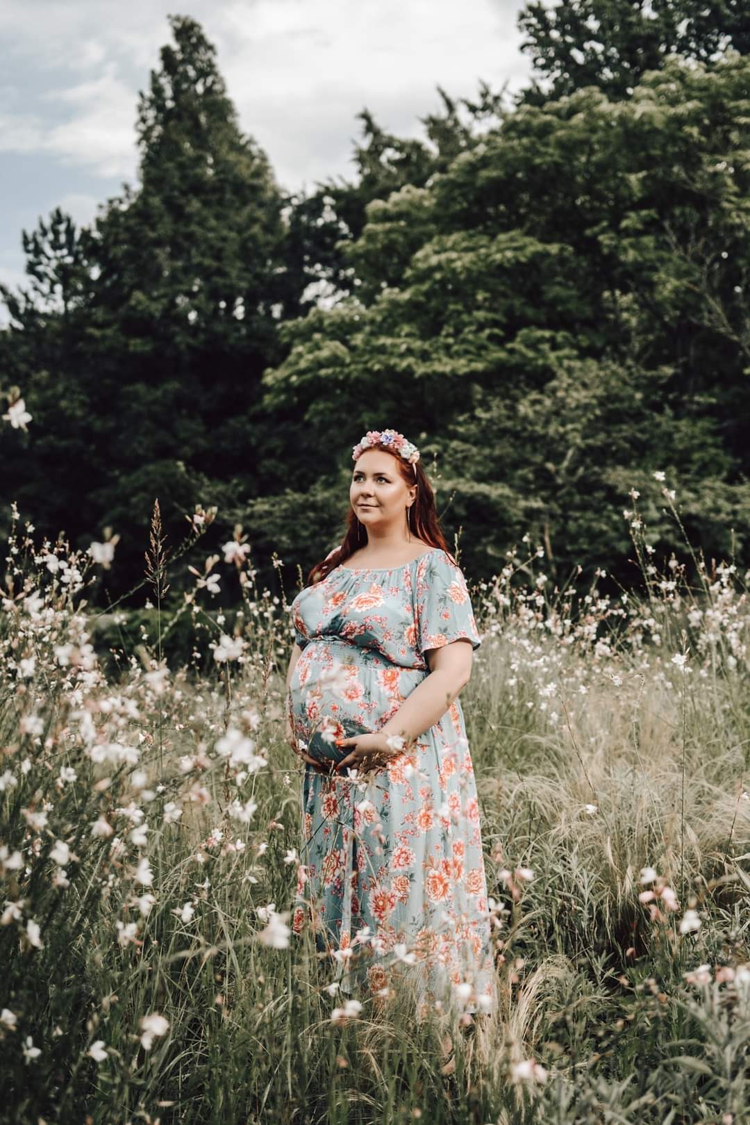 Kobieta w ciąży w kwiecistej sukience, stojąca na łące, z dłońmi na brzuchu i wiankiem na głowie, patrzy w bok.