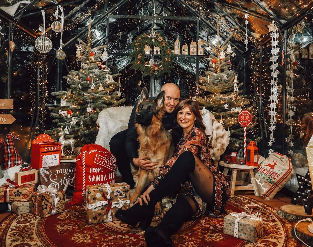 Uśmiechnięta para siedzi na dywanie z owczarkiem niemieckim w świątecznej scenerii szklarni, otoczona prezentami, choinkami z lampkami i dekoracjami.