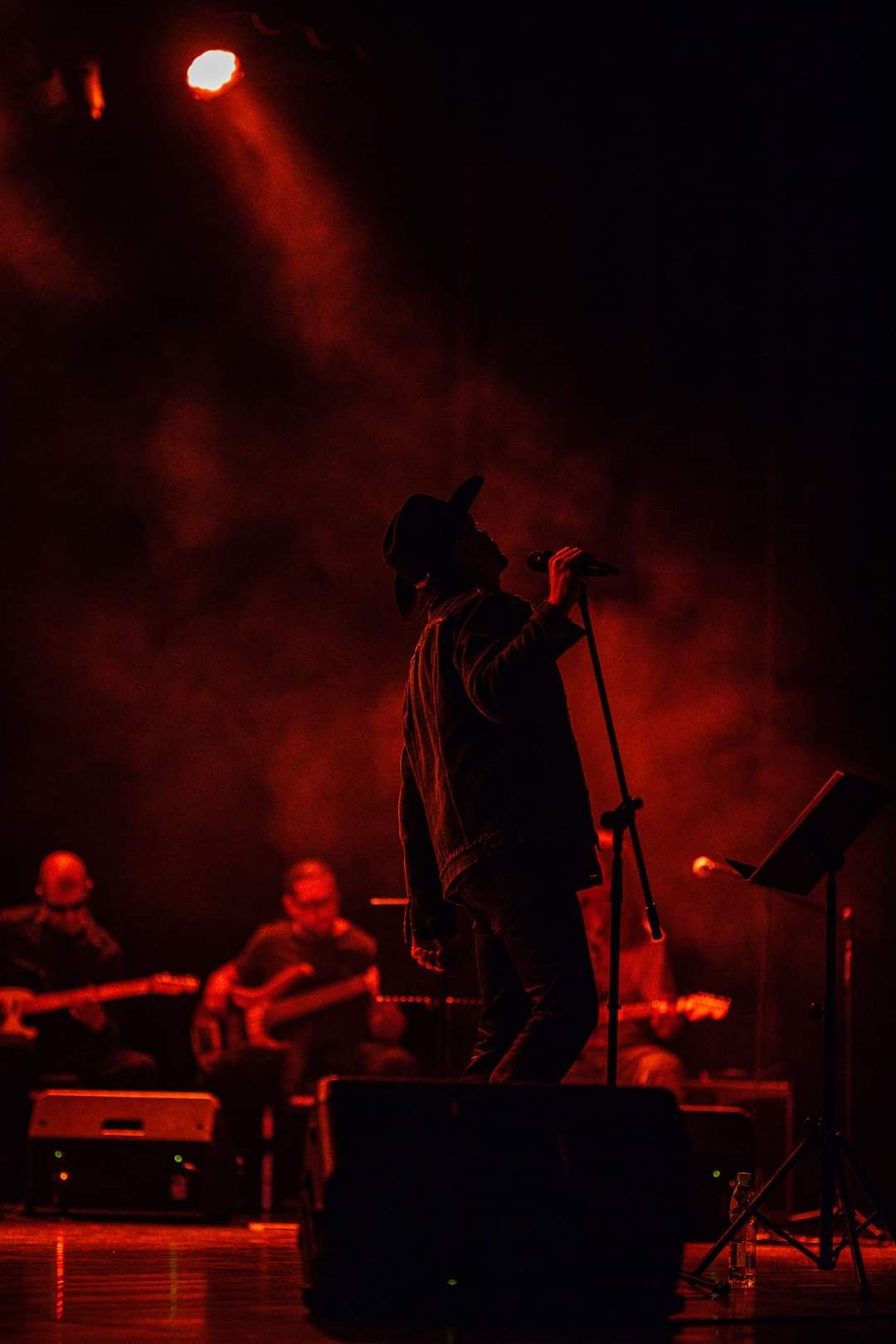 Sylwetka piosenkarza w kapeluszu śpiewającego do mikrofonu na scenie oświetlonej czerwonym światłem, z zespołem muzycznym w tle.