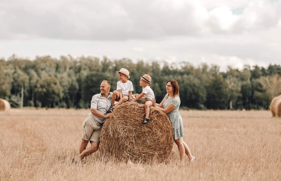 Rodzinne zdjęcie na tle pola ze słomianymi balami: ojciec i dwaj synowie siedzą na balocie, matka stoi obok, wszyscy uśmiechnięci, w tle las i pochmurne niebo.