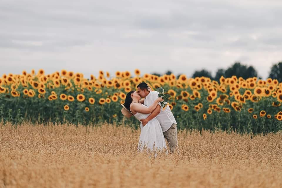 Zakochana para całuje się na polu zboża z kwitnącymi słonecznikami w tle, kobieta w białej sukience i kapeluszu, mężczyzna w białej koszuli i beżowych spodniach trzyma bukiet.