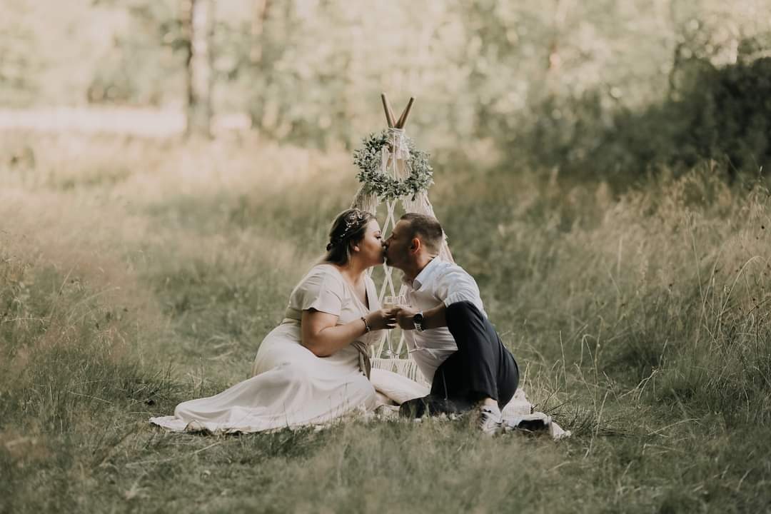Zakochana para całuje się na łące, siedząc przed małym tipi ozdobionym zielonym wieńcem, trzymając kieliszki z szampanem.