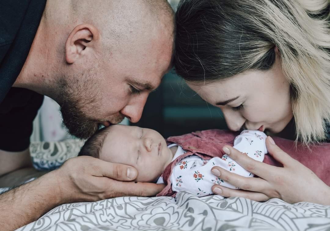 Rodzice delikatnie całują śpiące niemowlę w czoło, leżące na wzorzystej pościeli, widać fragment ubranka niemowlęcia w drobne kwiatki.
