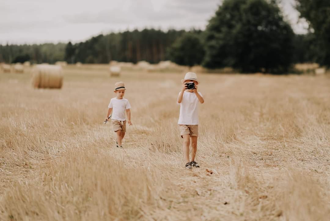 Dwóch chłopców w identycznych strojach i kapeluszach na polu ze słomą; jeden robi zdjęcie aparatem.