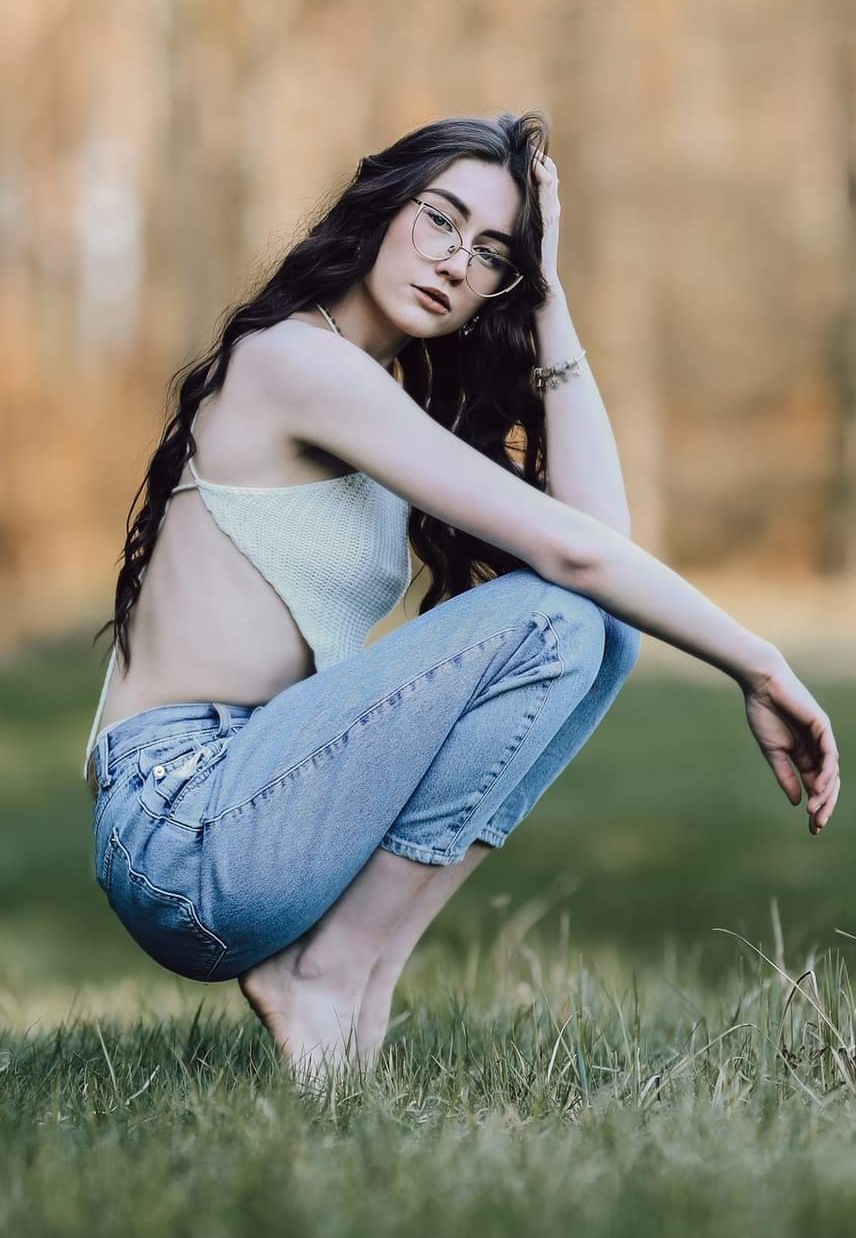 Portret kobiety w okularach pozującej w przysiadzie na trawie, ubrana w dżinsy i ażurowy top na tle rozmytej zieleni i brązu.