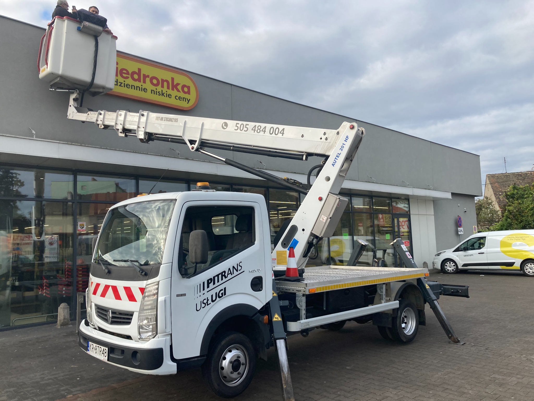 Biały samochód z wysięgnikiem na parkingu przed sklepem. W koszu wysięgnika dwóch mężczyzn. Na wysięgniku widoczny numer telefonu. Na samochodzie logo firmy Pitrans Usługi.