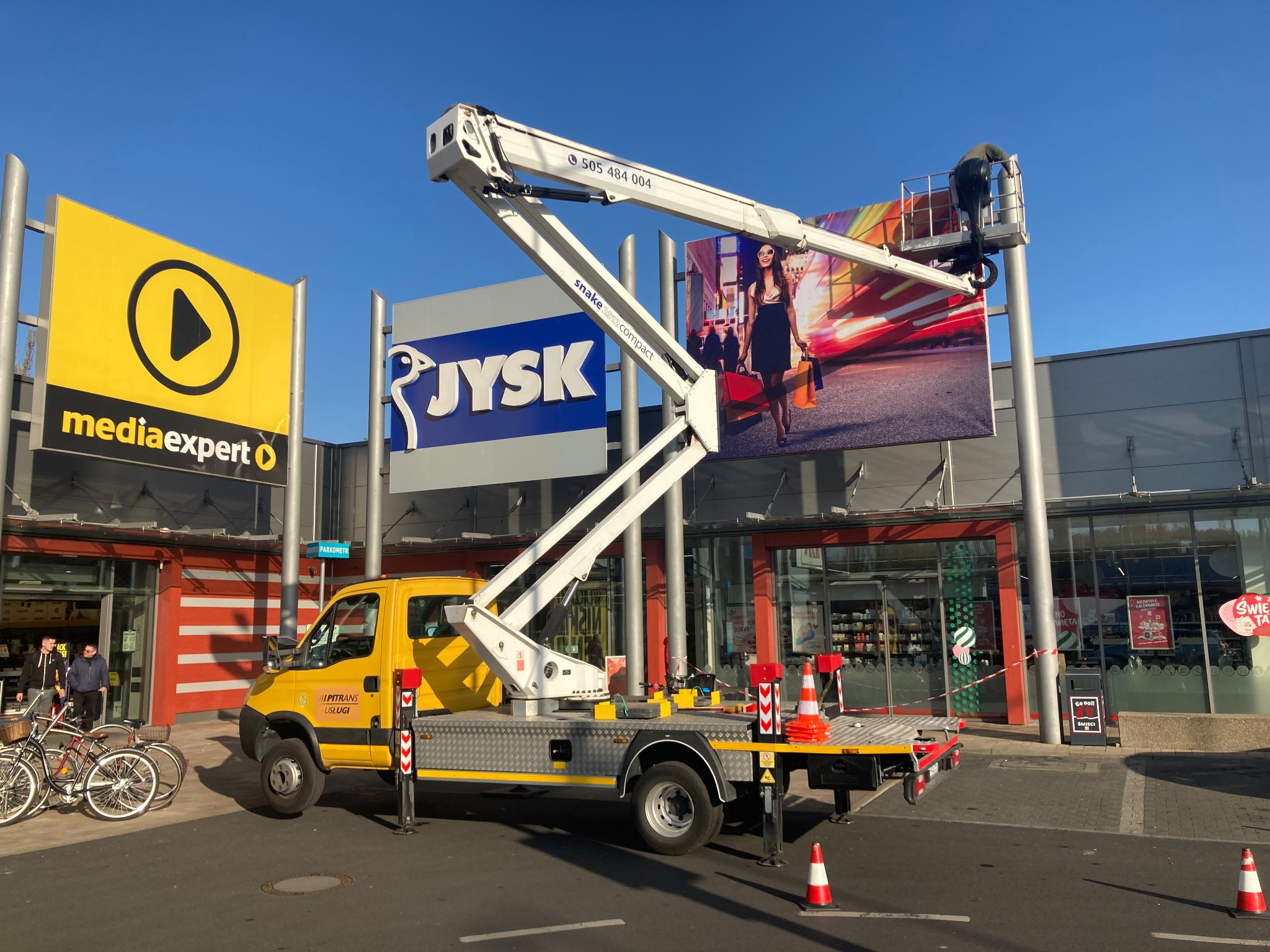 Żółty pojazd ze specjalistycznym wysięgnikiem podnosi pracownika do montażu lub naprawy dużego billboardu reklamowego przed sklepami Media Expert i JYSK w słoneczny dzień.