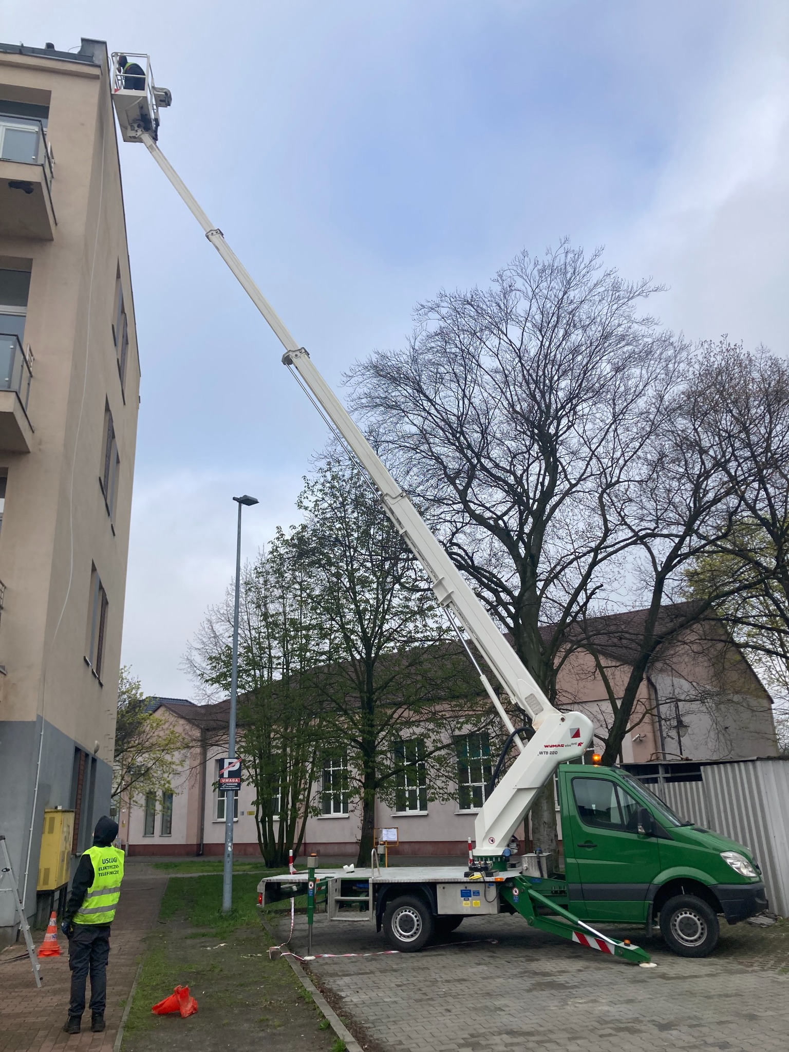 Zielony samochód z podnośnikiem koszowym wysięgnikiem przy budynku mieszkalnym, pracownik w żółtej kamizelce obserwuje z dołu.