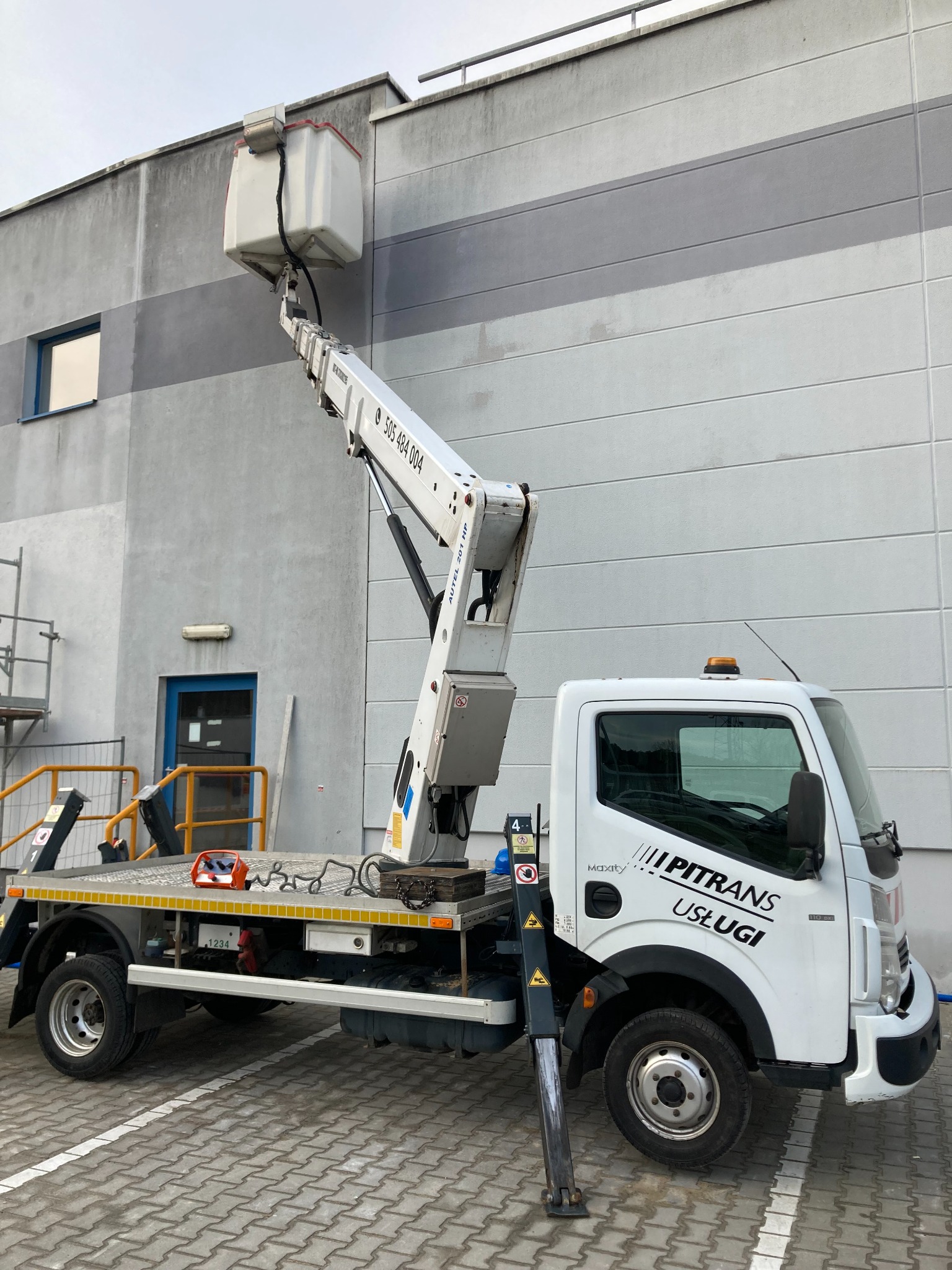 Biały podnośnik koszowy firmy z Zielonej Góry, z wysięgnikiem ustawionym przy szarej ścianie budynku, na kostce brukowej.