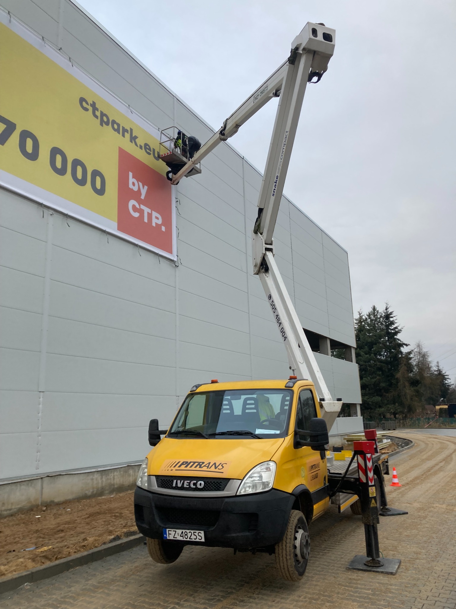 Żółty podnośnik koszowy Iveco Pitrans z wysięgnikiem Snake Compact na tle szarej hali, pracownik w koszu montuje duży baner reklamowy CTPark.eu.