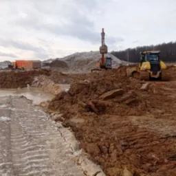 Żółty spychacz Komatsu i koparka na placu budowy z błotnistym terenem i śladami opon, w tle wywrotka i drzewa pod pochmurnym niebem.