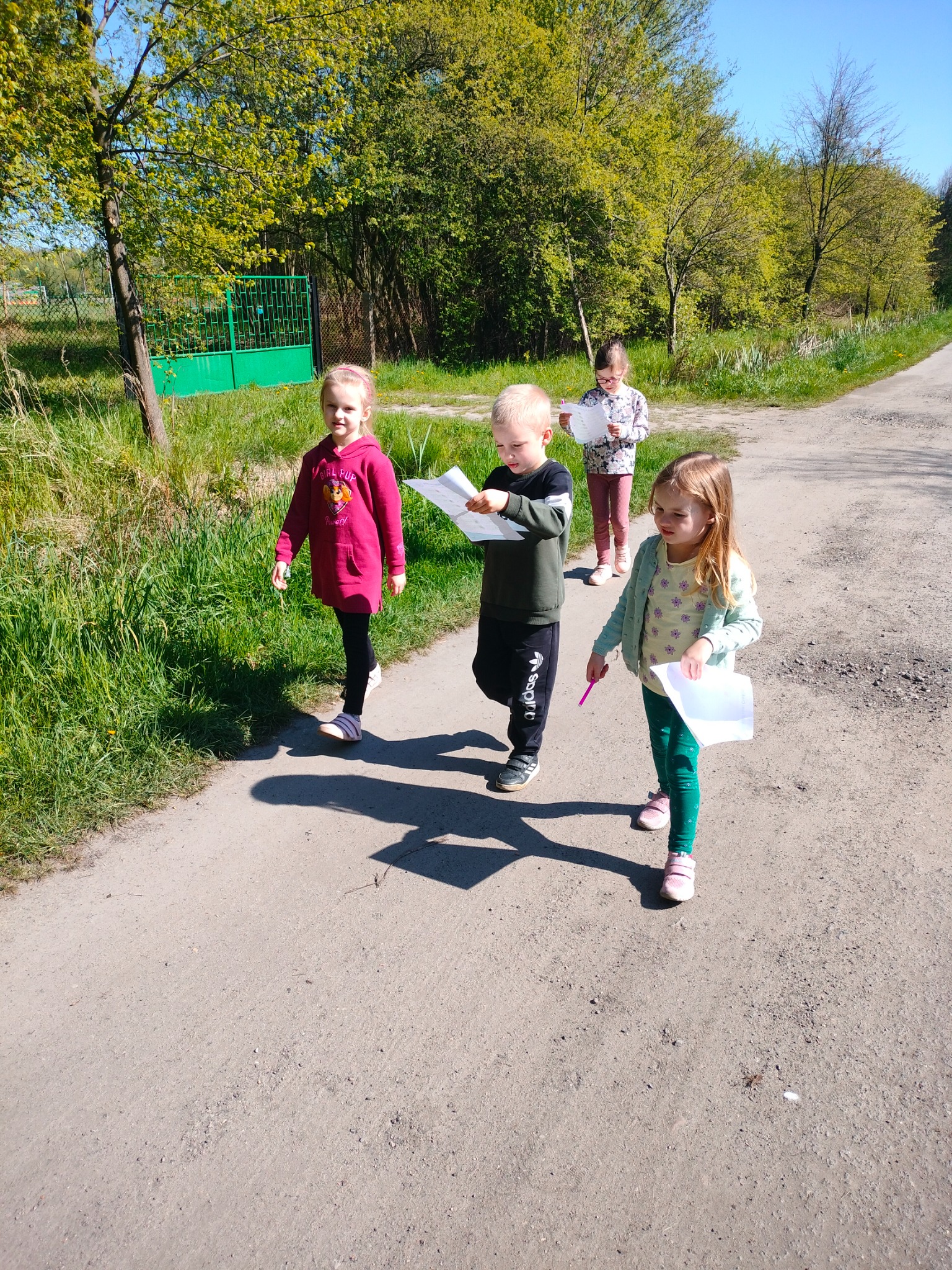 Czwórka dzieci idzie polną drogą, trzymając w rękach kartki papieru i długopis; prawdopodobnie w trakcie zajęć edukacyjnych na świeżym powietrzu.