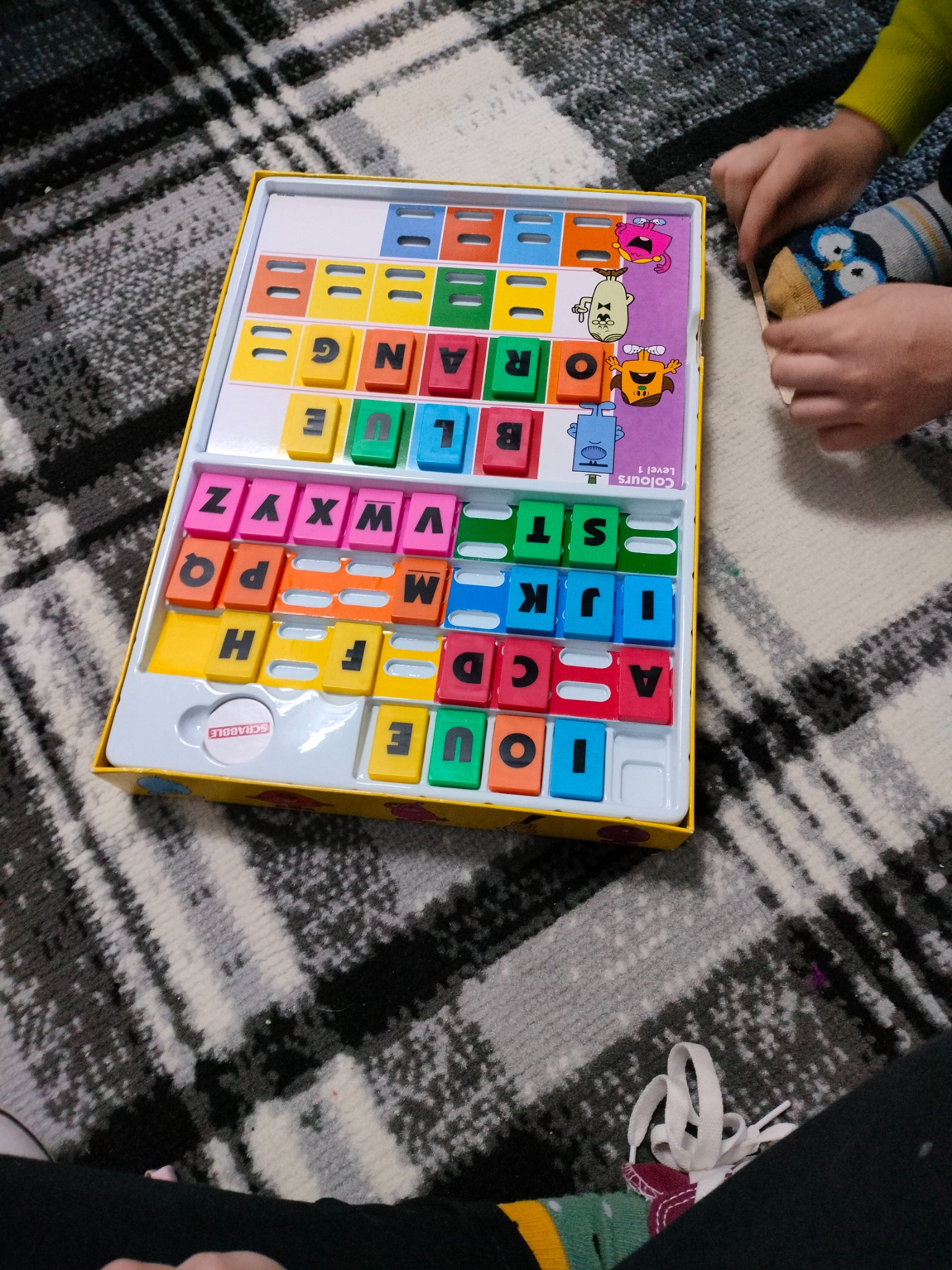 Dziecko w skarpetkach w pingwiny bawi się edukacyjną grą Scrabble Junior, ucząc się kolorów i słów, na dywanie w geometryczne wzory.