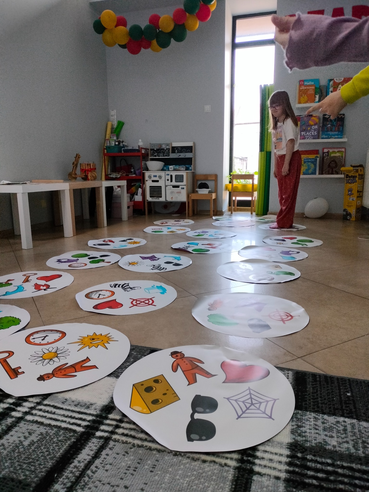 Dziewczynka w okularach stoi na kartce z obrazkiem podczas zabawy edukacyjnej w sali z dekoracjami.