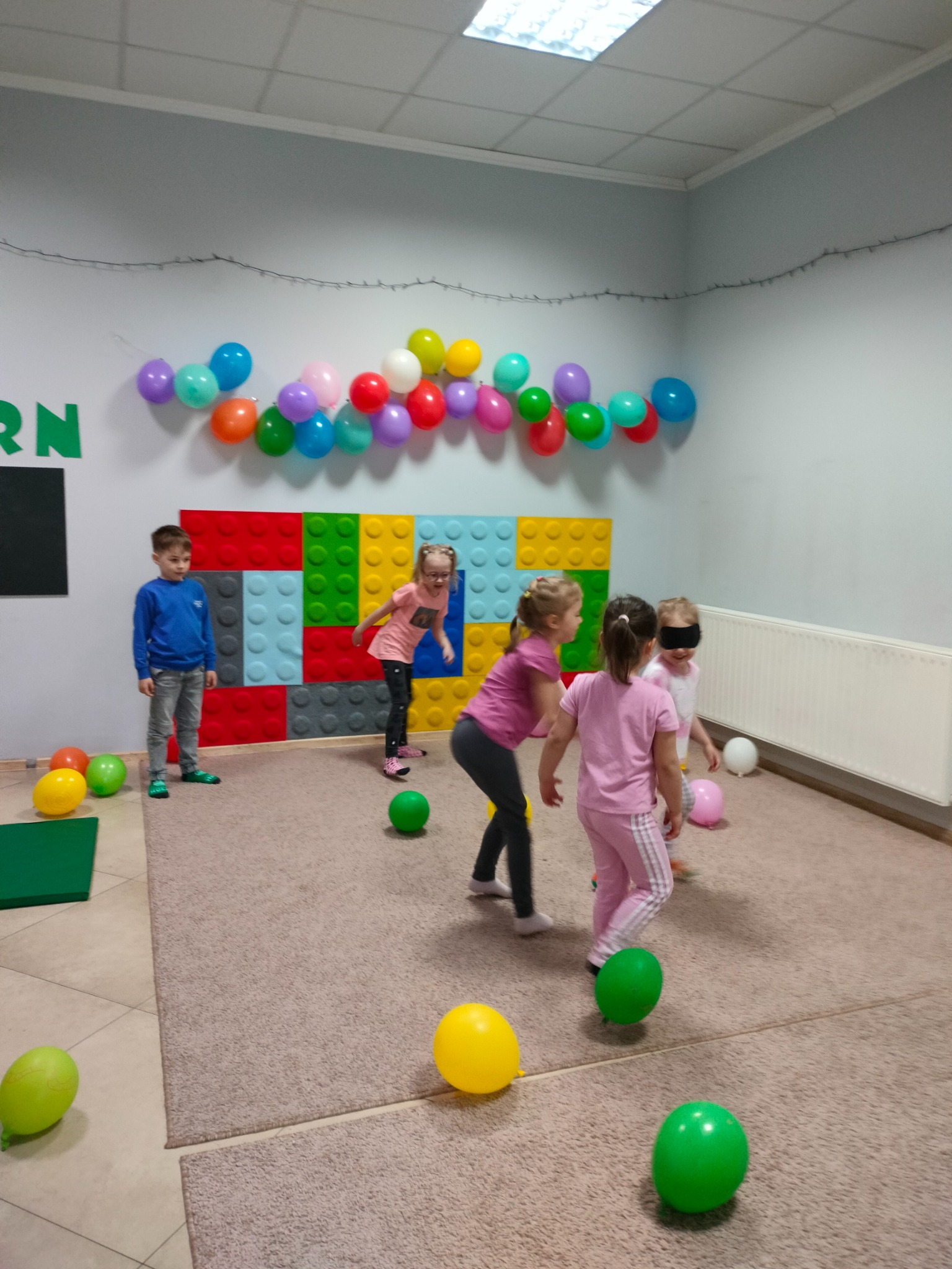 Grupa dzieci w jasnym pokoju bawi się w grę z balonami, jedna dziewczynka ma zasłonięte oczy, na ścianie dekoracja z balonów i kolorowe panele przypominające klocki Lego.