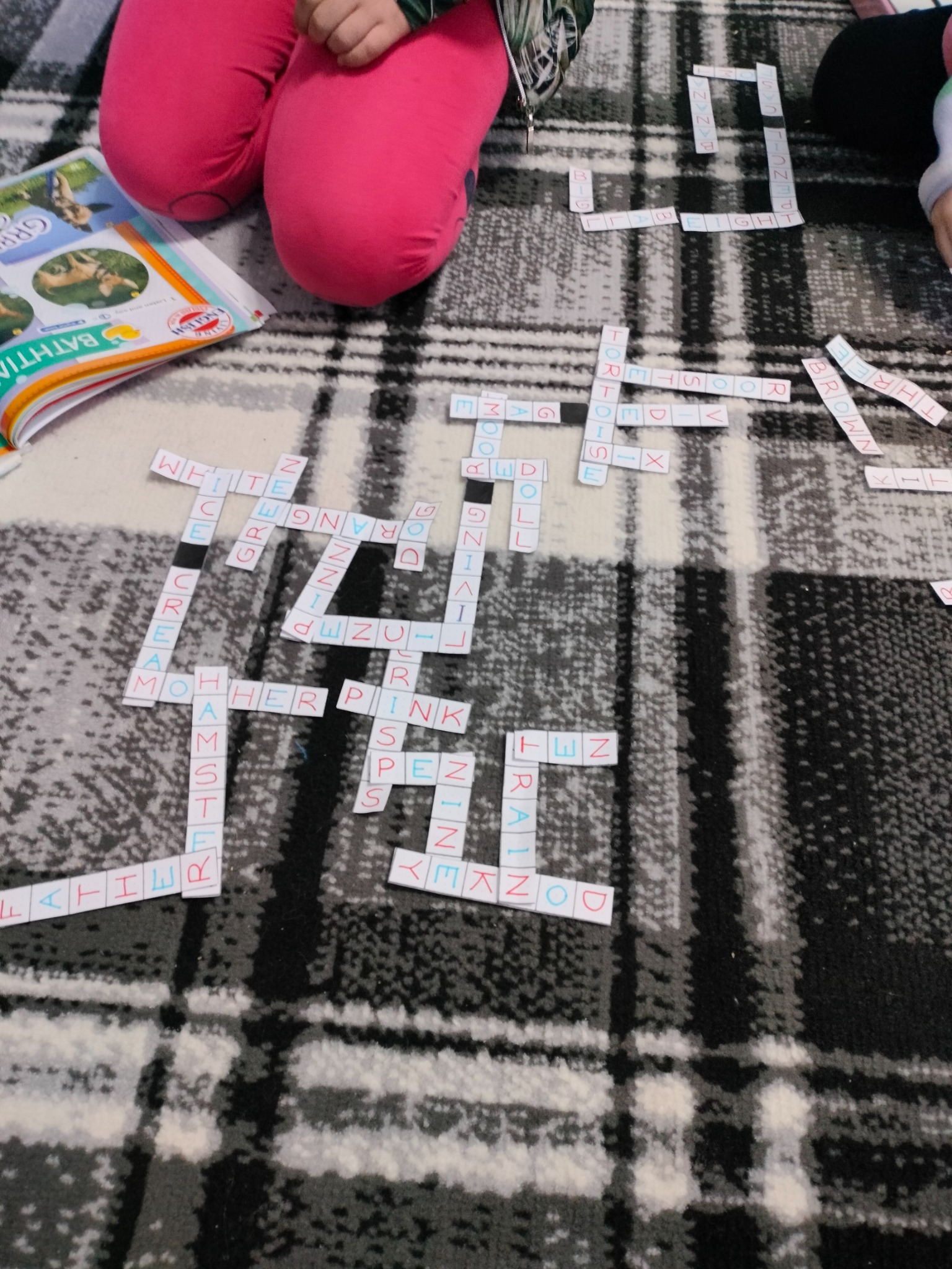 Dziecko układa słowa z kartoników na kocu w kratę. Widoczne słowa to między innymi: WHITE, ORANGE, PINK, FATHER, BROWN, BANANA, ENGLISH.