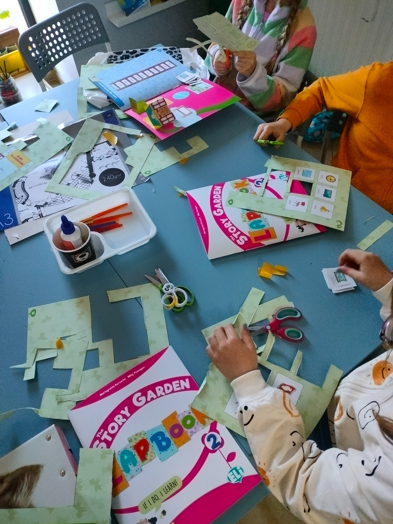 Dzieci wycinają elementy z papieru do interaktywnej książki 'The Story Garden Lapbook 2', na niebieskim stole rozłożone nożyczki, klej i kolorowe paski papieru.