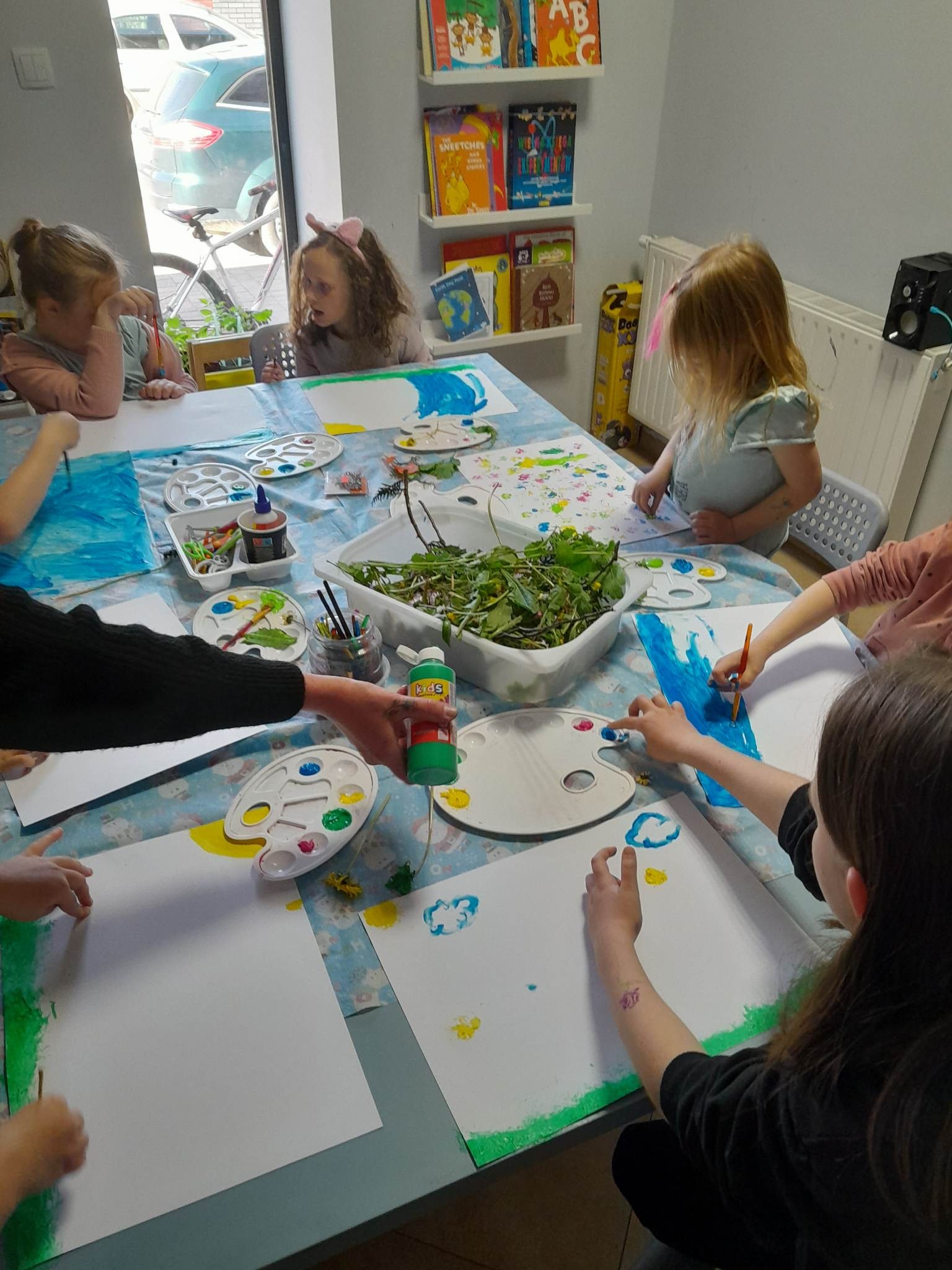 Dzieci podczas zajęć plastycznych, malowanie farbami na papierze, używanie naturalnych elementów jak liście do stemplowania, widoczne palety z farbami, pędzle i pojemnik z zielonymi roślinami.