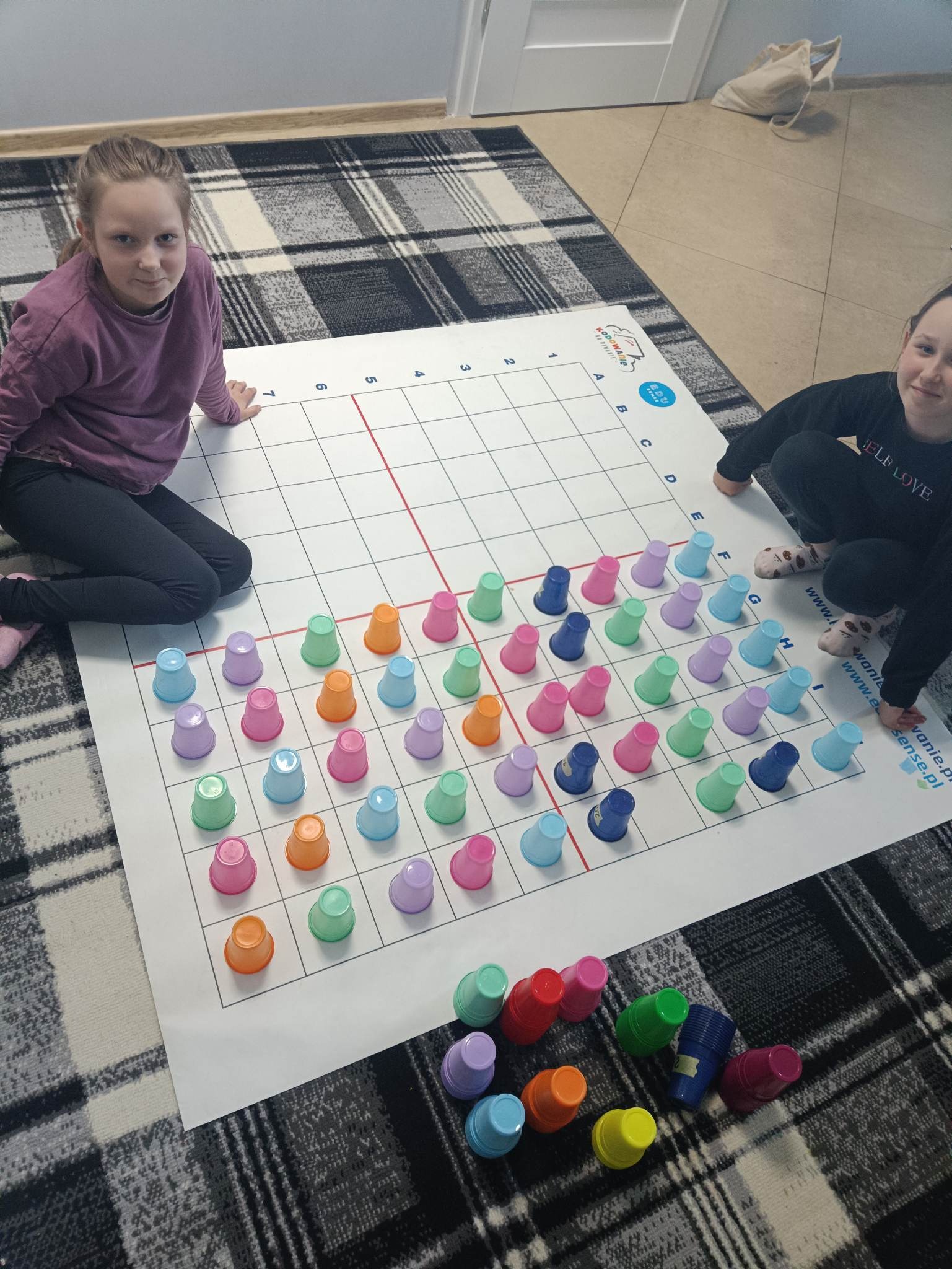 Dwie dziewczynki siedzą na podłodze obok dużej planszy z siatką i kolorowymi kubeczkami, układając je według określonego wzoru, prawdopodobnie w ramach zajęć edukacyjnych z kodowania.