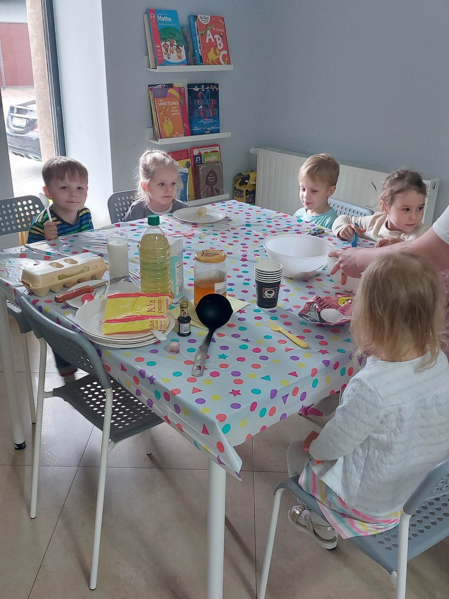 Grupa dzieci siedzi przy stole z kolorowym obrusem, przygotowując składniki do pieczenia, widoczne jajka, mąka, olej, miód i miska, a w tle półka z książkami dla dzieci.