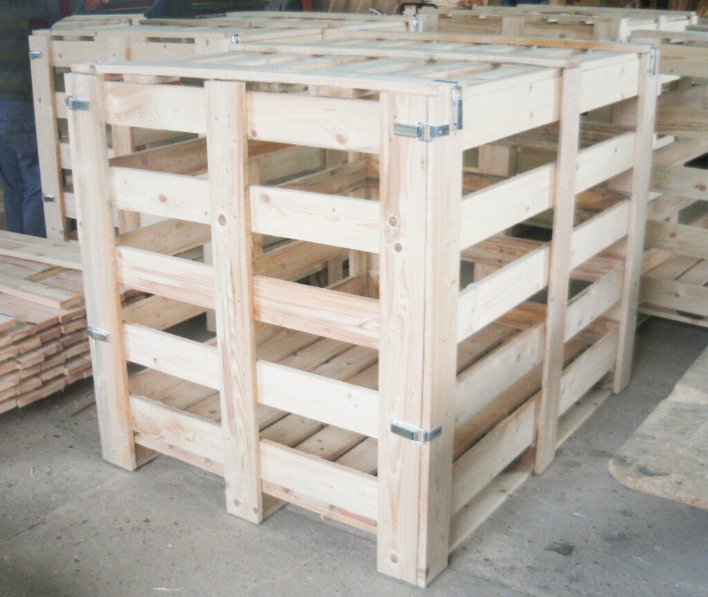 Drewniana, ażurowa skrzynia transportowa z jasnego drewna, zabezpieczona metalowymi klamrami, ustawiona na betonowej posadzce, w tle widoczne inne skrzynie i stos desek.